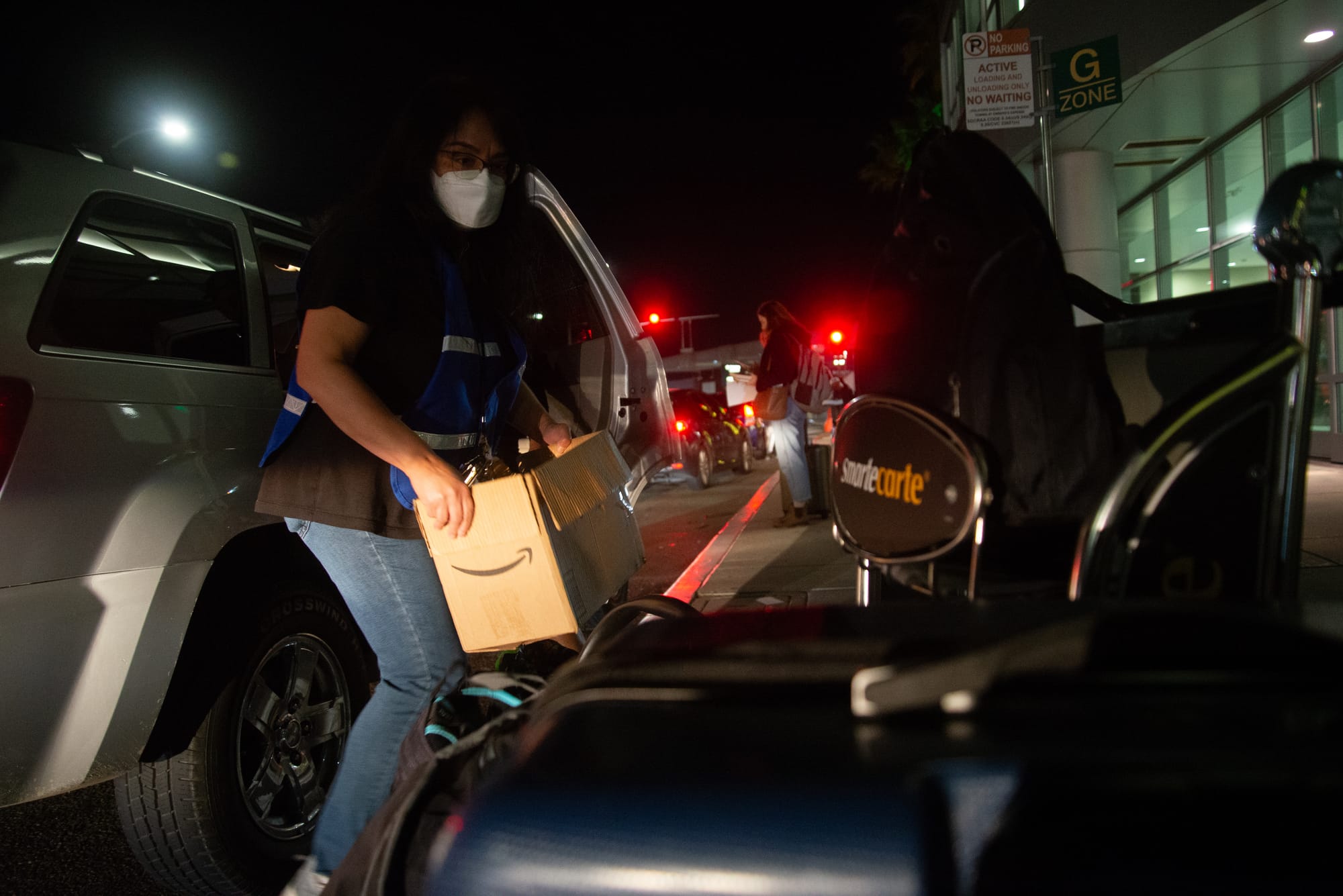 A woman picks up a box between a car and a luggage cart outside the airport