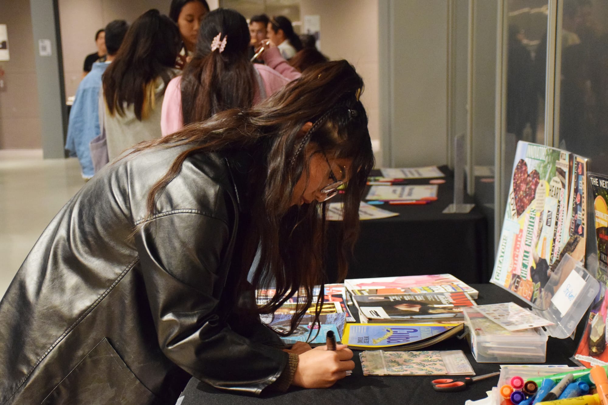 A person with long hair and a leather jacket draws on a craft table as people gather in groups behind her at an event