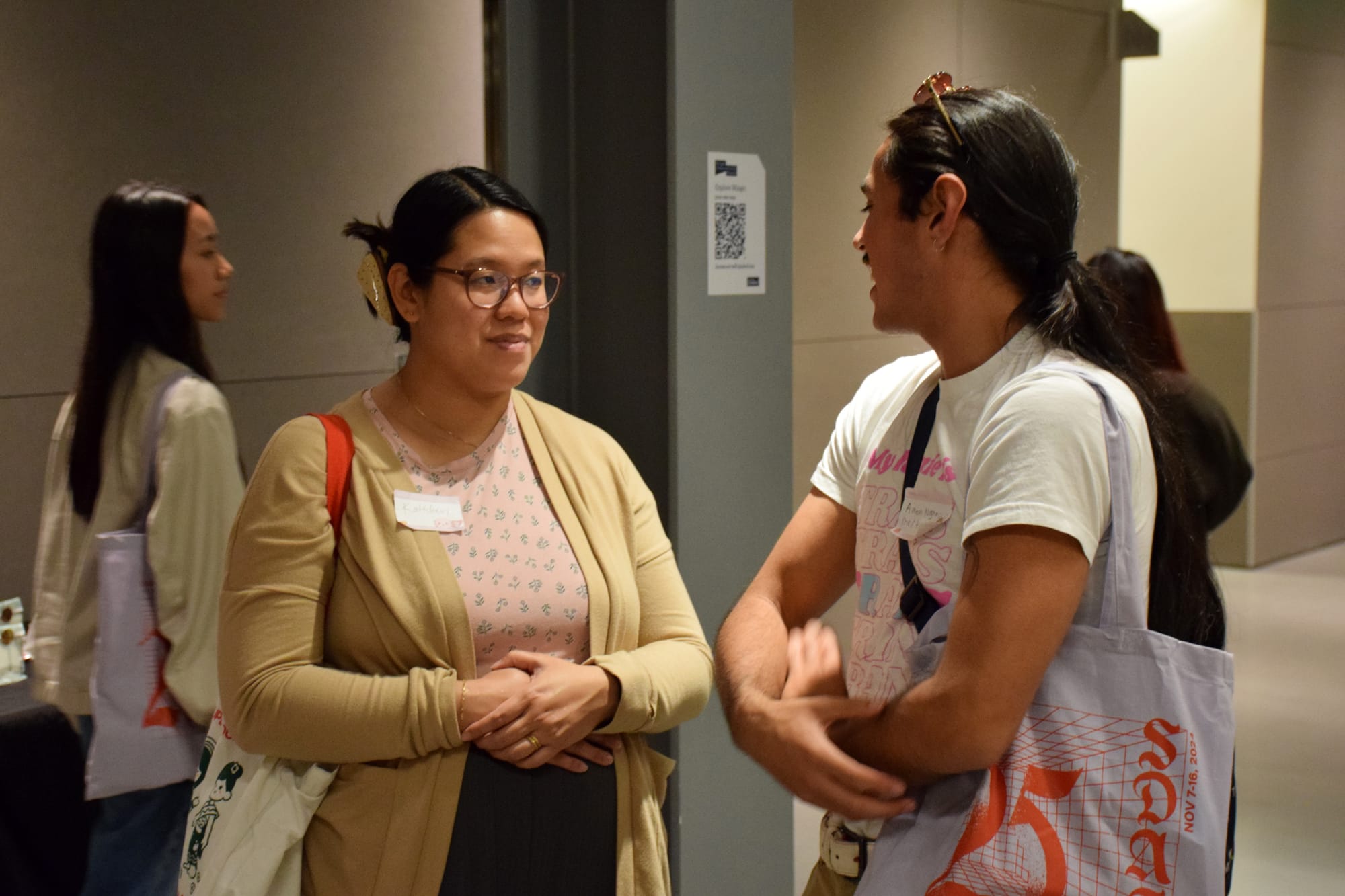 Two people speak to one another in a room with other people walking behind them