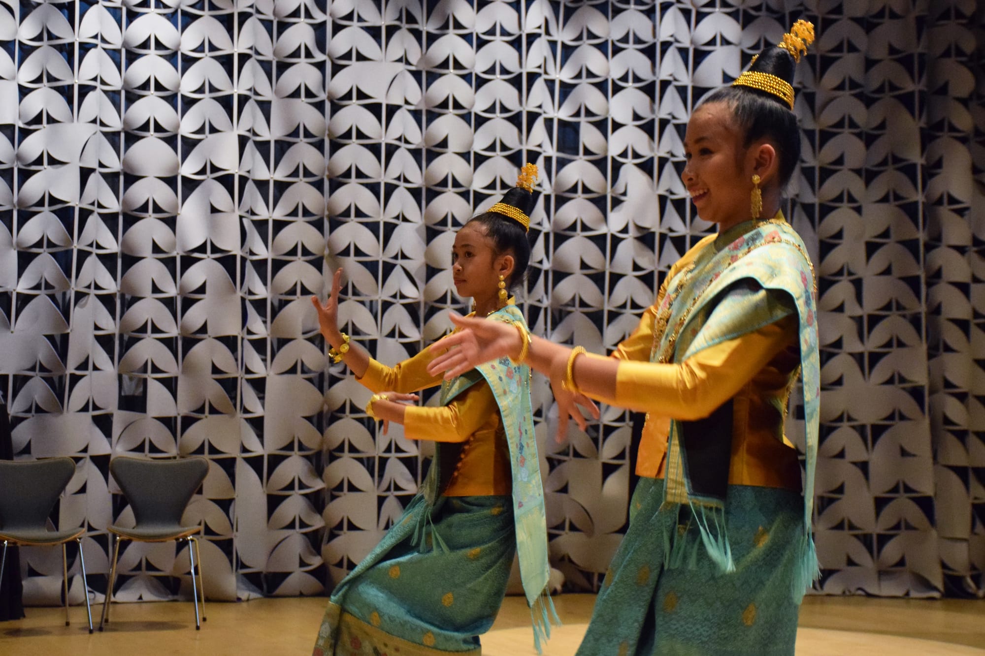 Two girls wearing traditional Laotian clothing and head pieces dance in front of a patterned curtain in a room