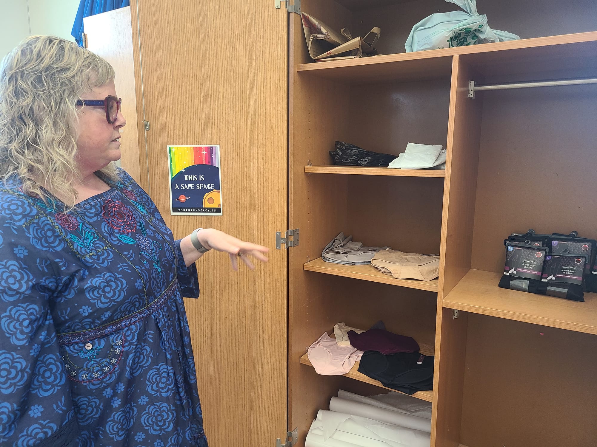 A woman in a blue floral dress stands in front of a classroom storage closet that has shelves of underwear, leggings and plastic bags.