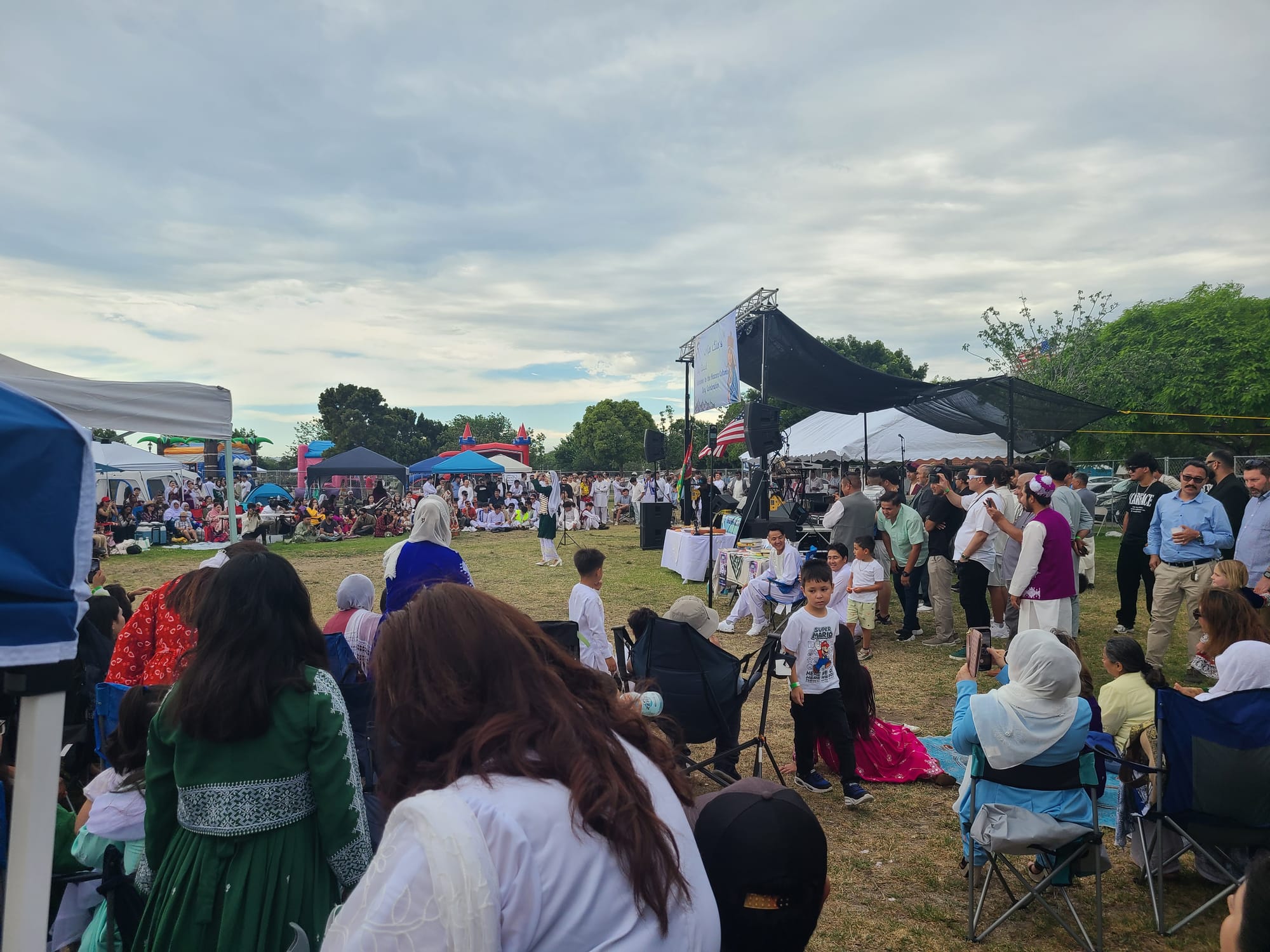 A crowd of people surround a grassy area in front of a stage where a woman in a green and white outfit dances a traditional Hazara dance