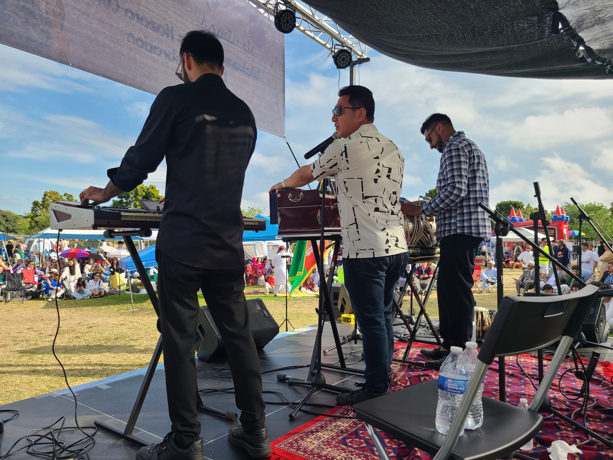 Three musicians, one at the keyboard, one with tablas and one with a harmonium stand on stage facing away from the camera