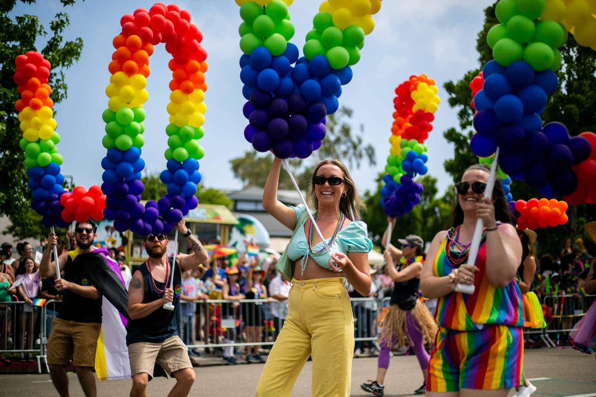 people in colorful outfits holding rainbow balloons walk in an outdoor parade in front of a crowd
