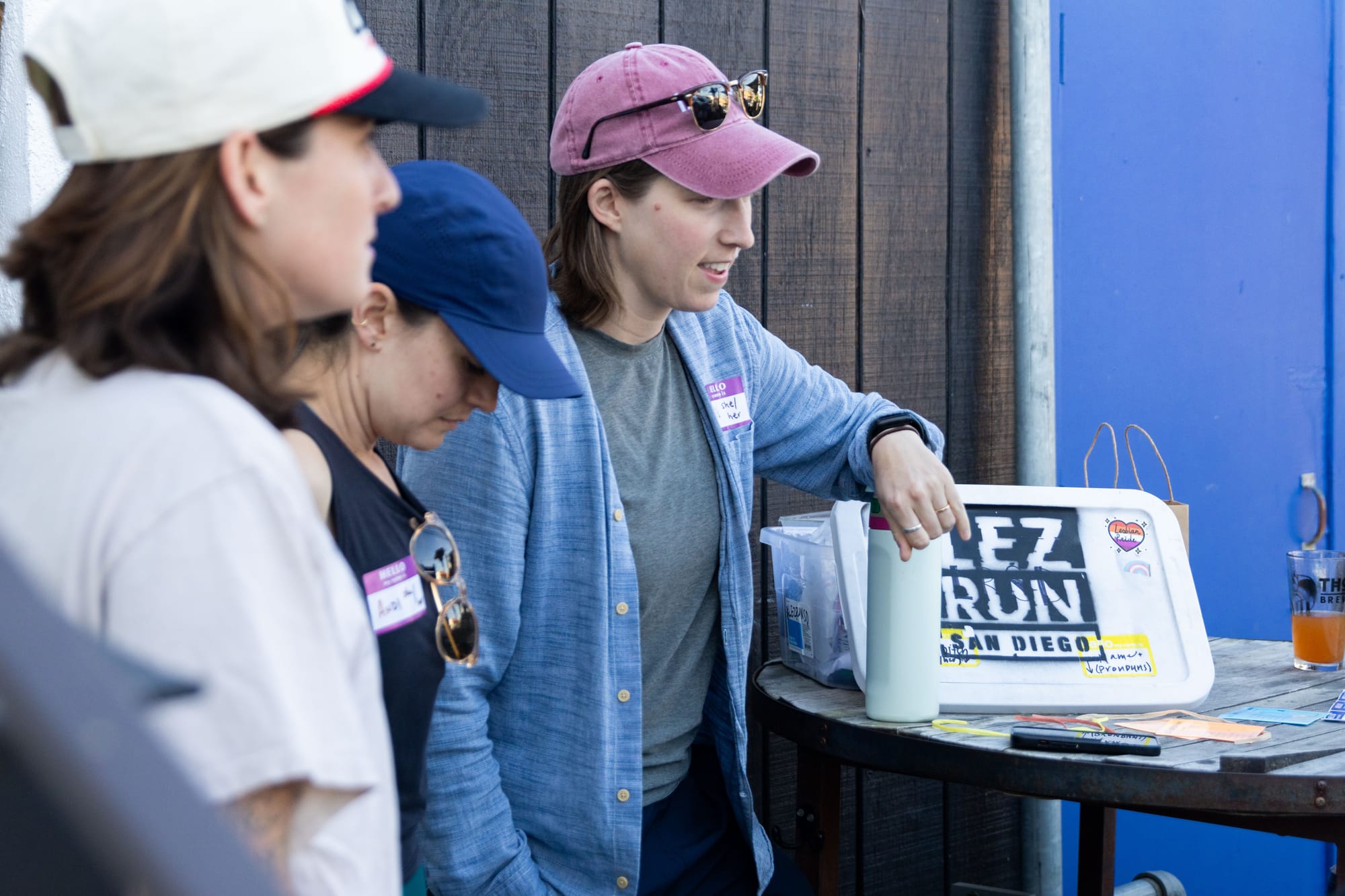 Three people stand outside next to a table with a plastic organizer box on it painted with the words "LEZ Run San Diego"