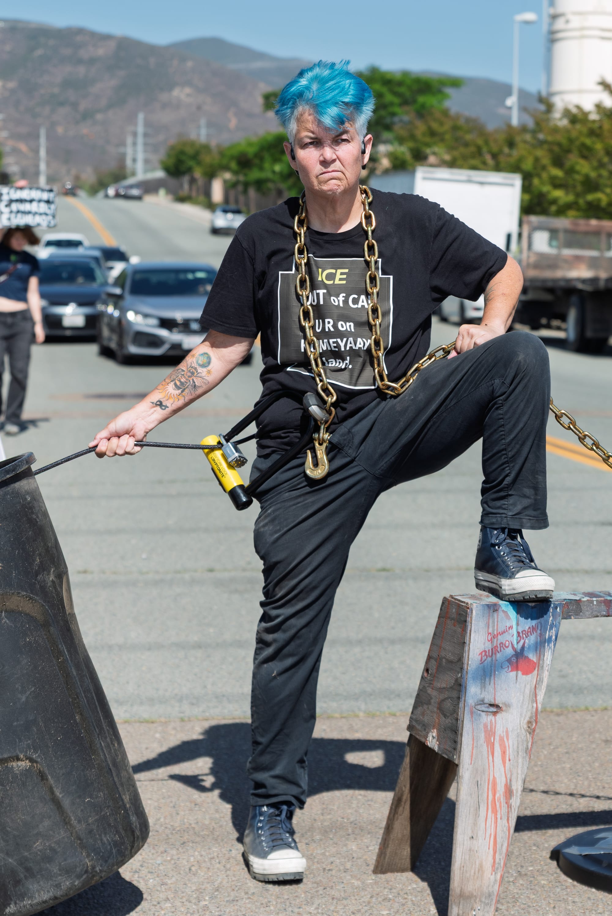 A woman with short, blue hair stands chained to trash cans in the street