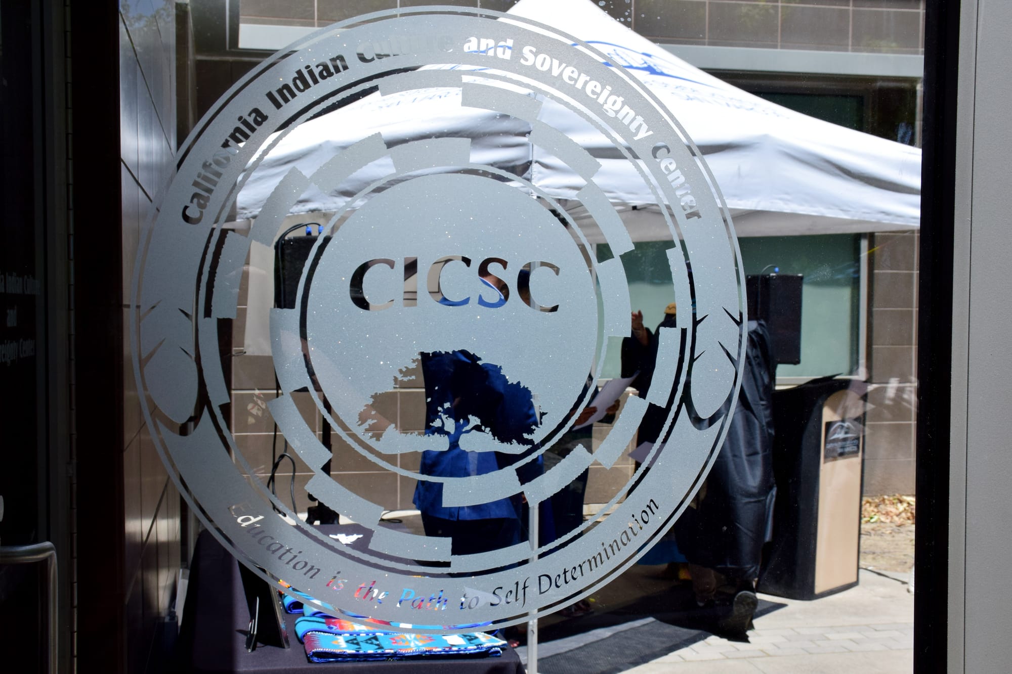 A photo of a frosted glass emblem on a door that reads CICSC: California Indian Culture Sovereignty Center, dedication is the path to self determination.