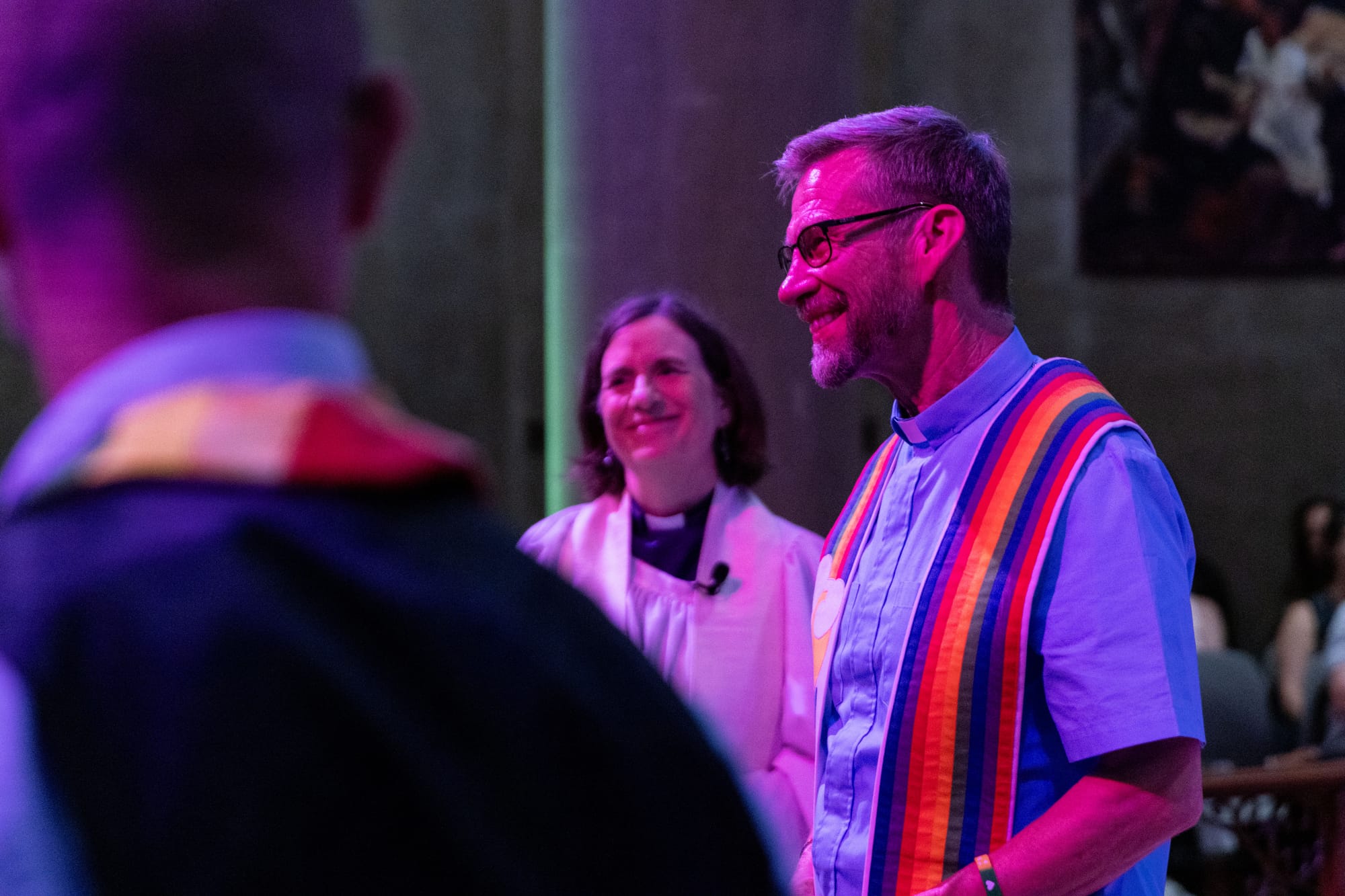 two pastors stand in a church, one in white robes and the other with a rainbow stole