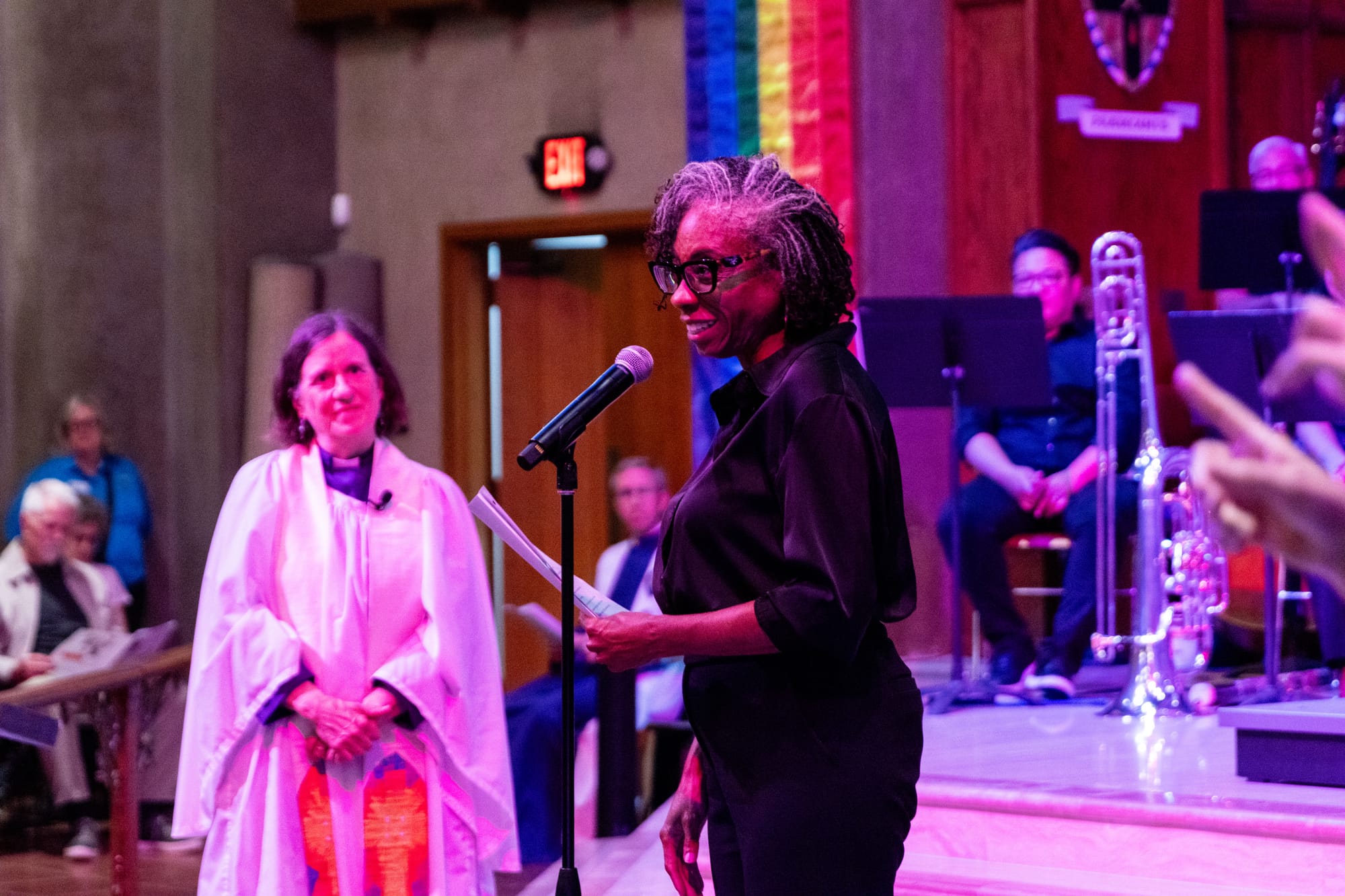 A woman speaks at a church in front of a symphony and a pastor in a white robe