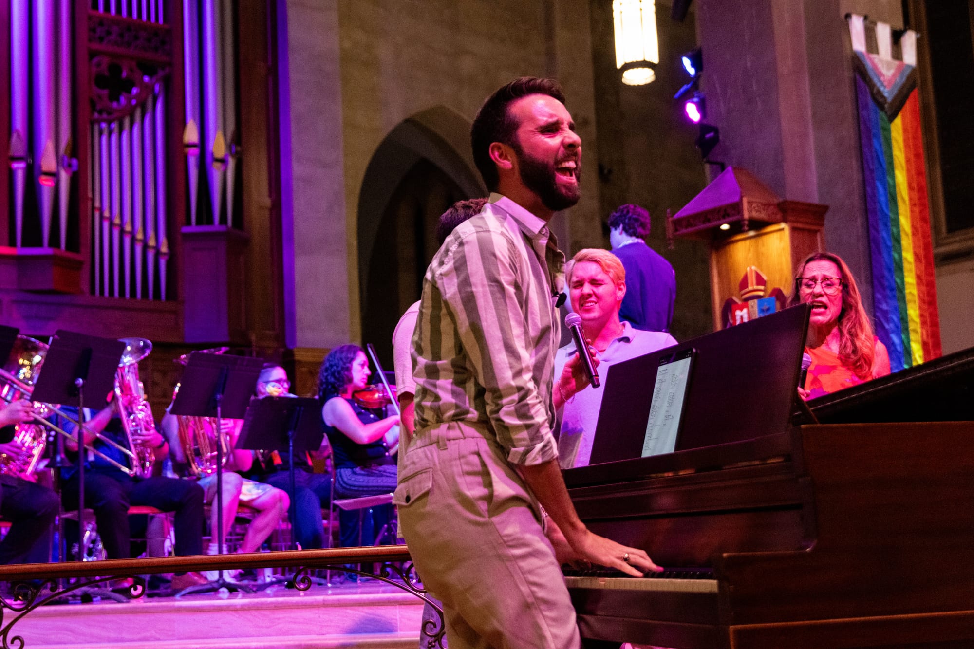 A person plays the piano in a church in front of a symphony, singers and a rainbow flag.