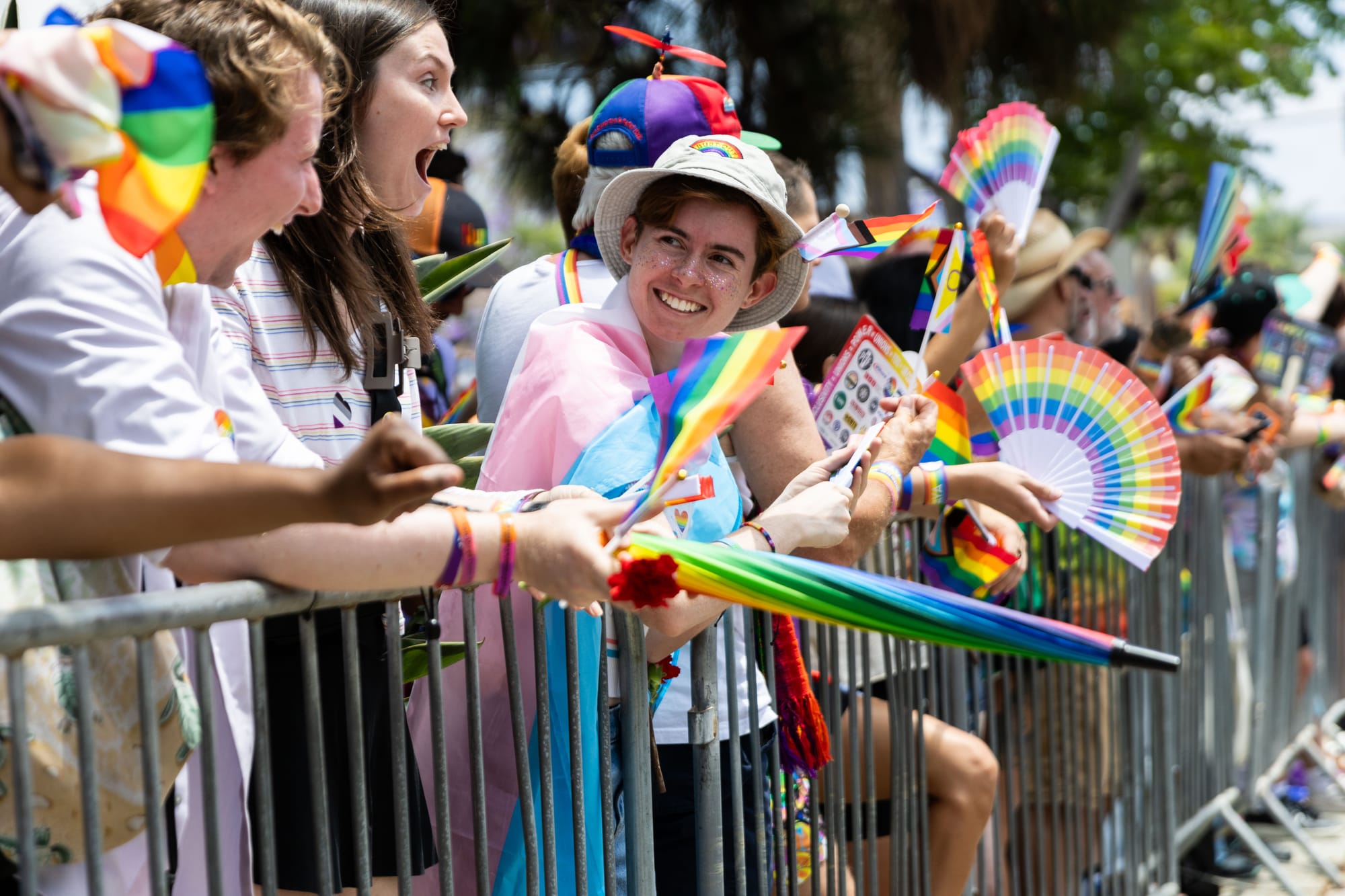 People stand next to a fence barrier holding rainbow flags, fans and other paraphernalia. 