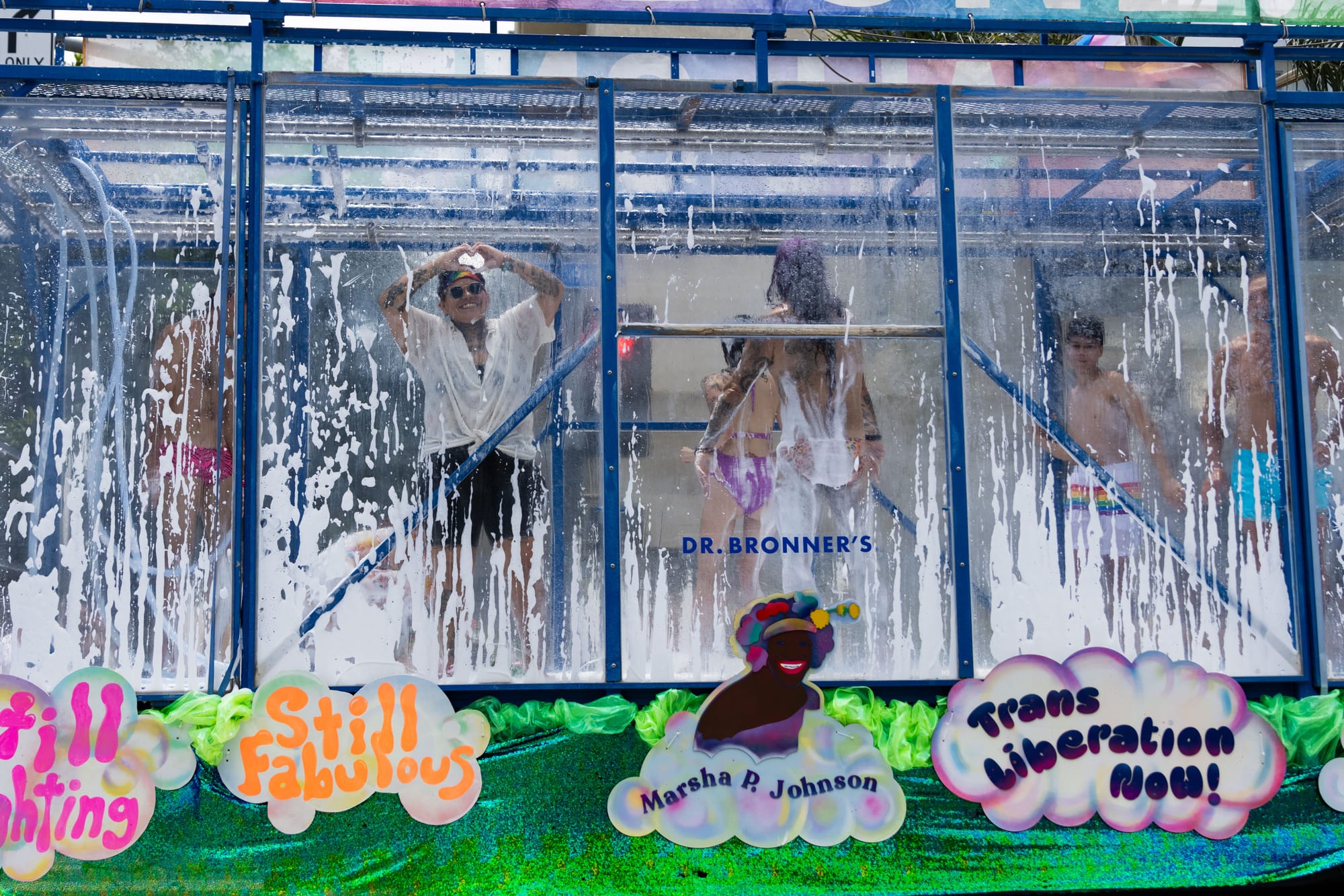 People dance in a glass parade float with foam spraying around inside. The float says "Still Fighting, Still Fabulous. Marsha P. Johnson. Trans Liberation Now!"