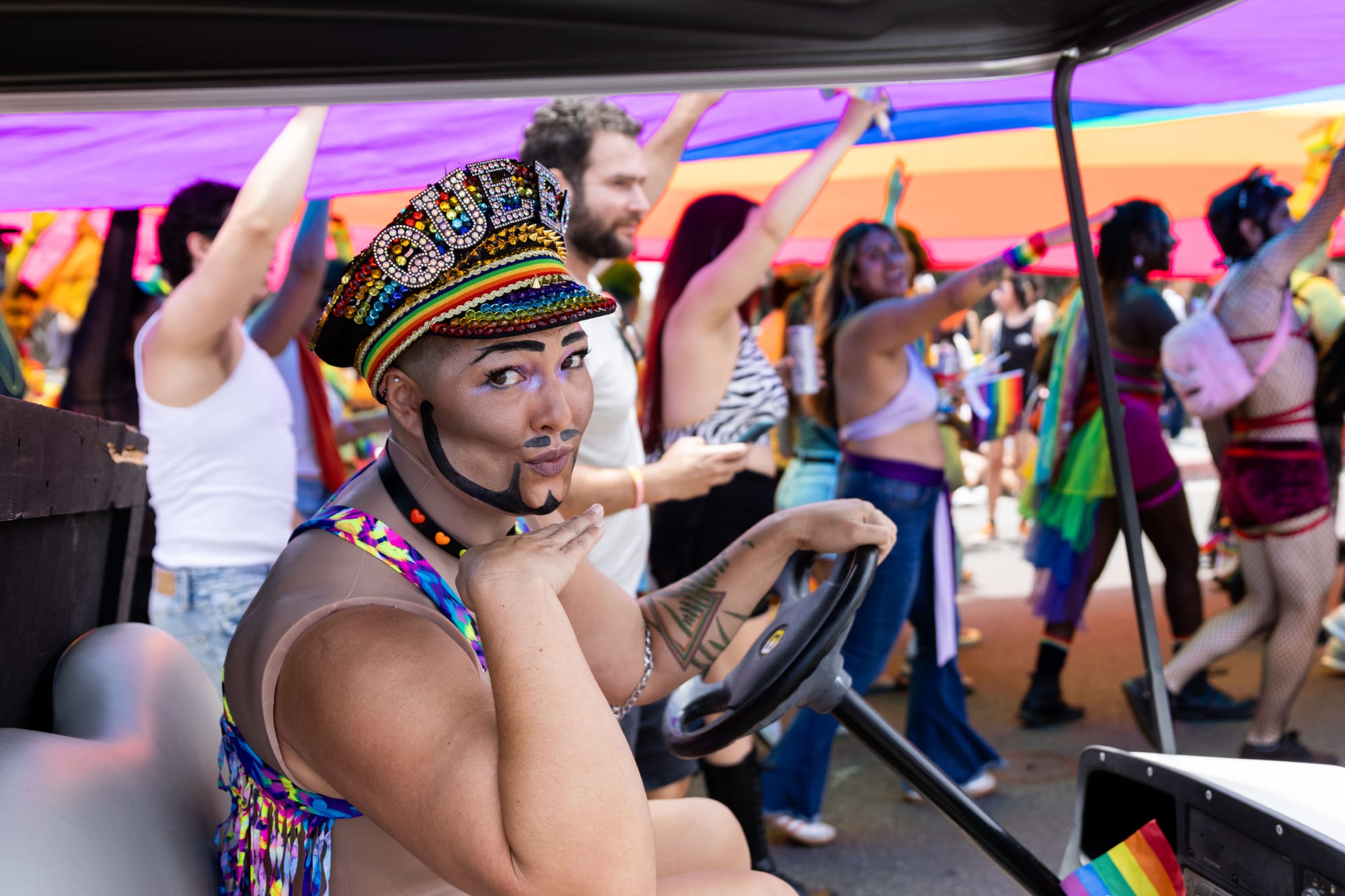 A person in drag with a fake beard and mustache drives a golf cart and poses in front of a crowd