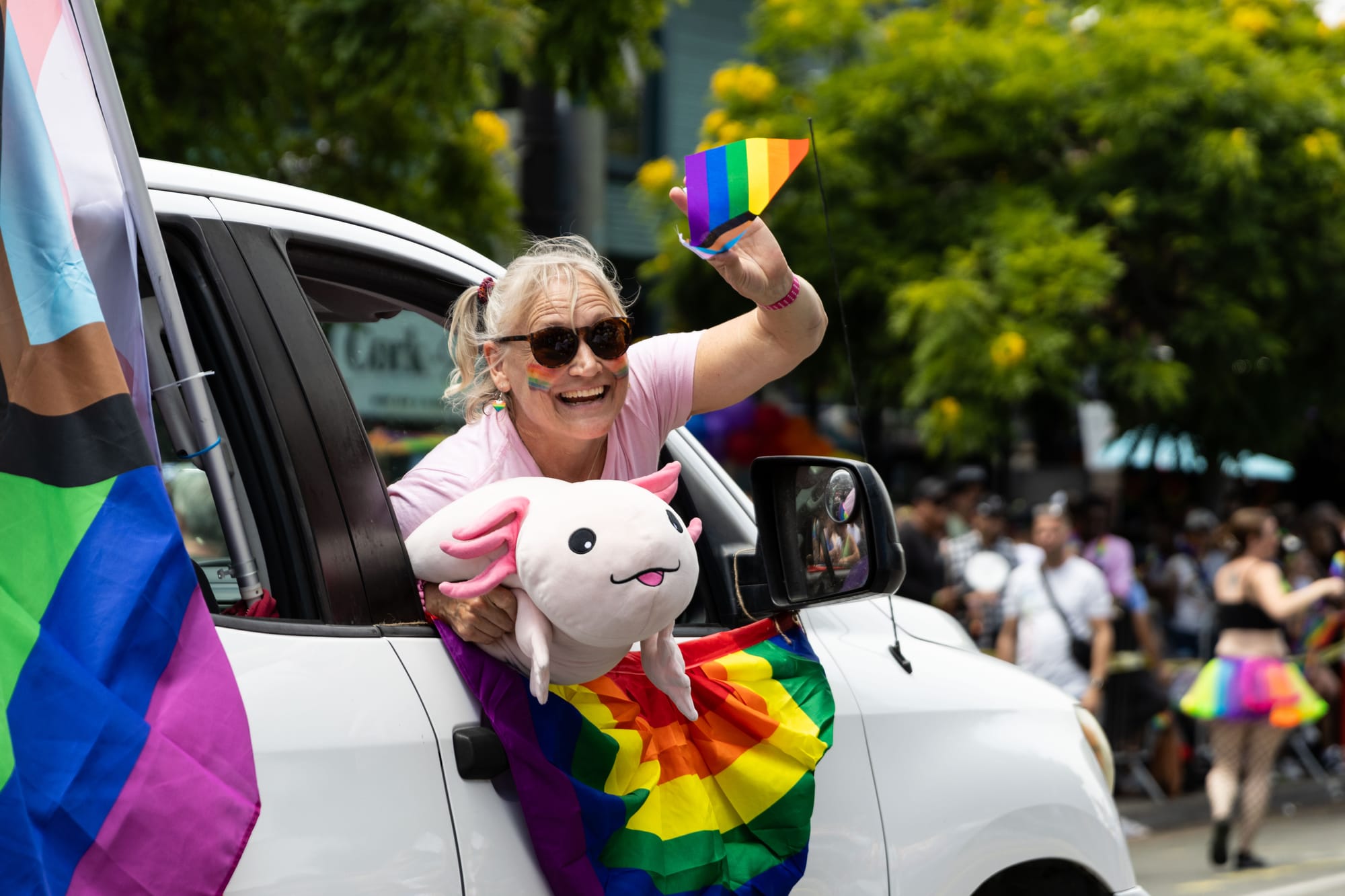Someone holds a stuffed axolotl toy and waves a rainbow flag from a car in a parade