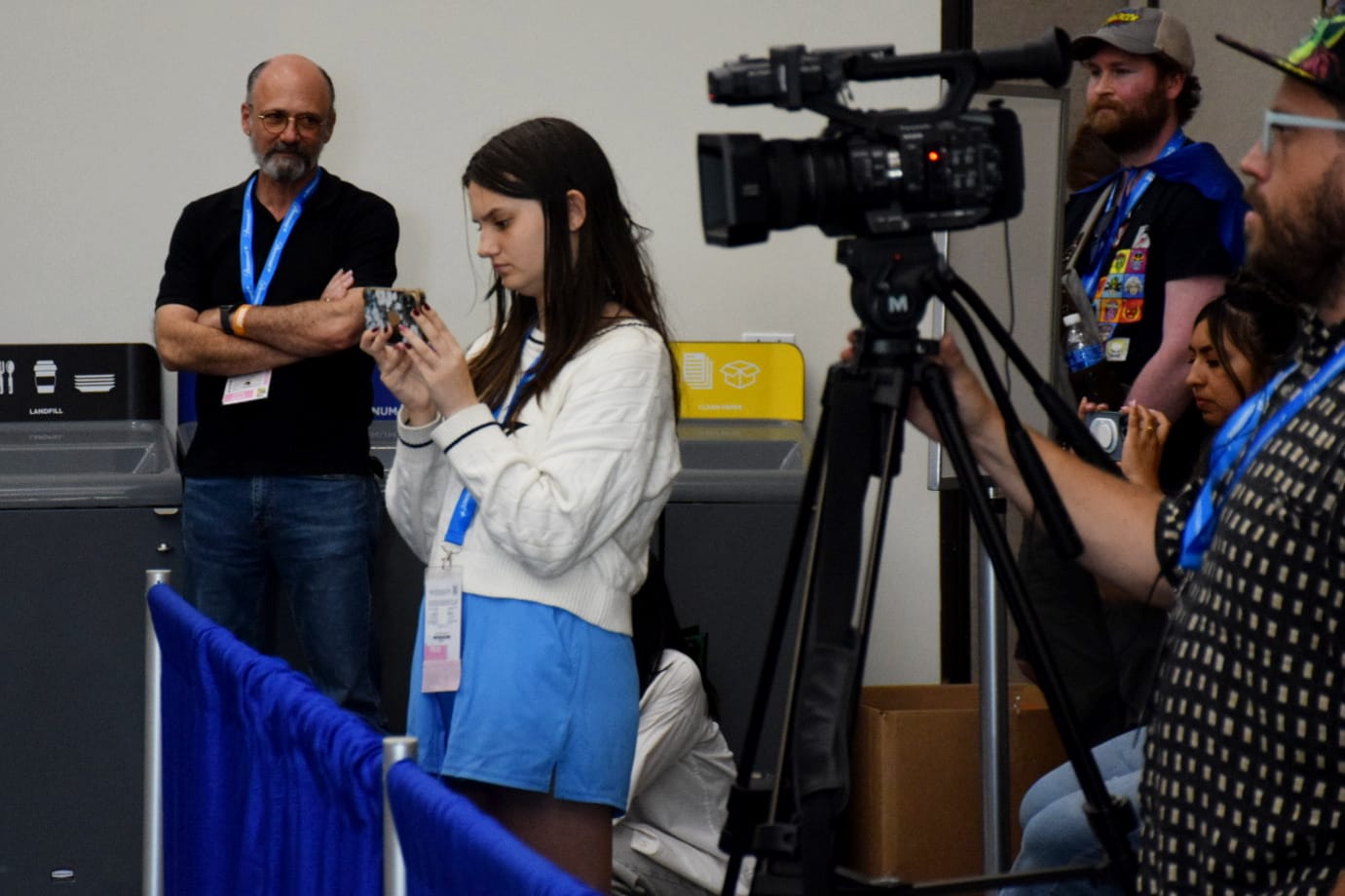 A teenager films something on her cell phone surrounded by other people, including a camera operator.