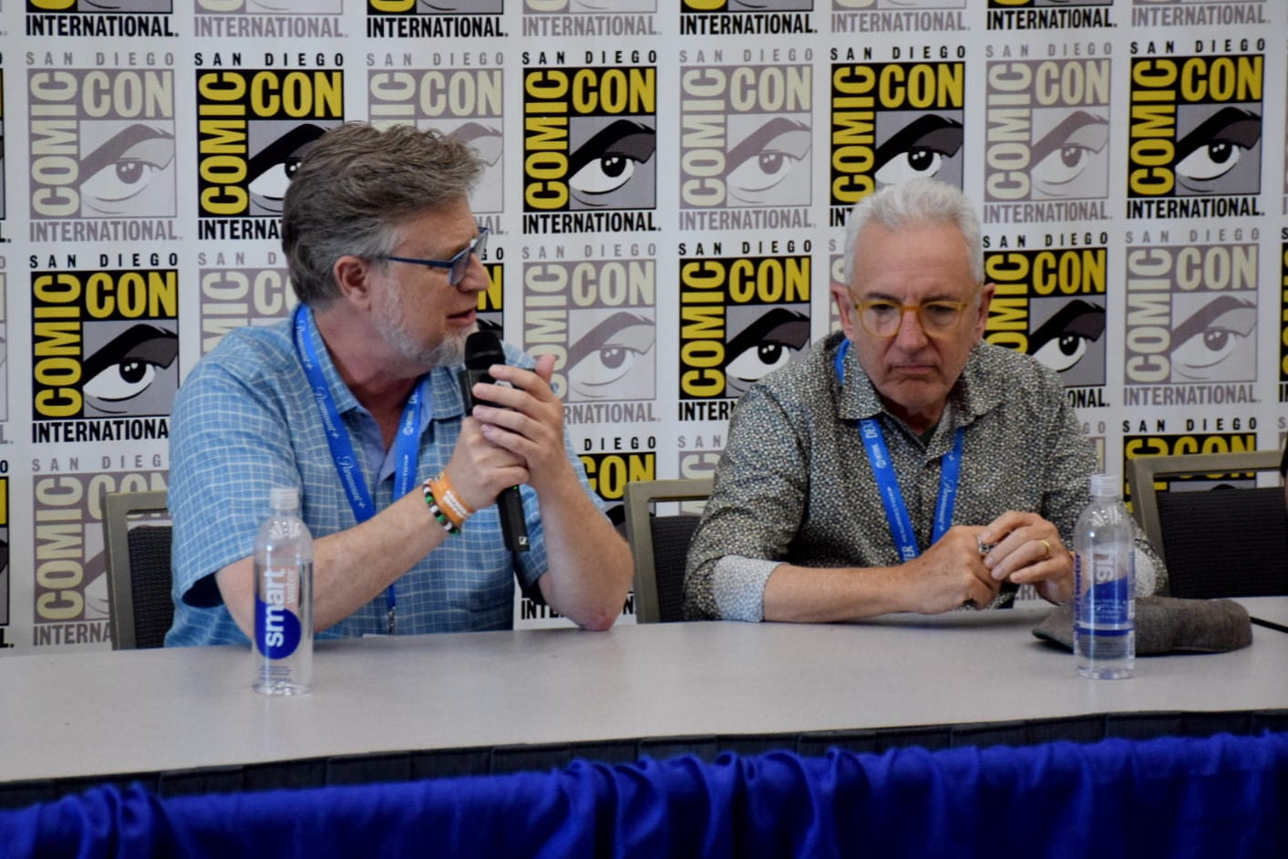 Two men sit at a panel table with the comic-con logo repeated on a backdrop behind them.
