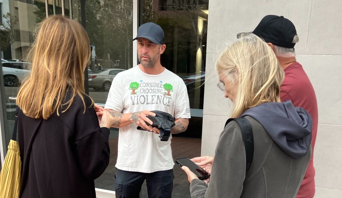 A man in a black cap and white shirt with the text consider choosing violence stands in talking with three people whose backs are toward the camera