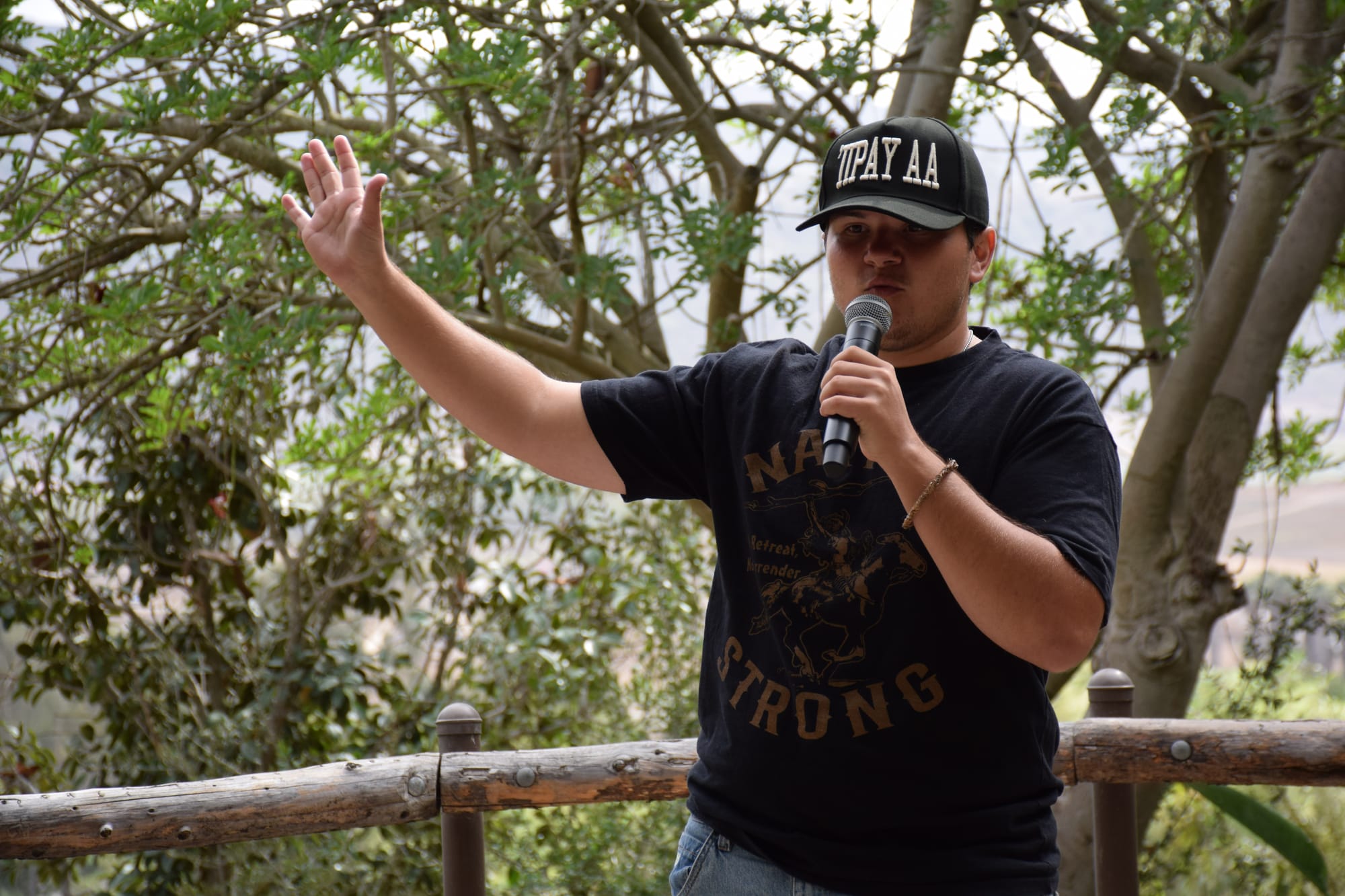 A person wearing a t-shirt that reads "Native Strong" and a hat reading "'Iipay AA" speaks into a microphone during an outdoor event.