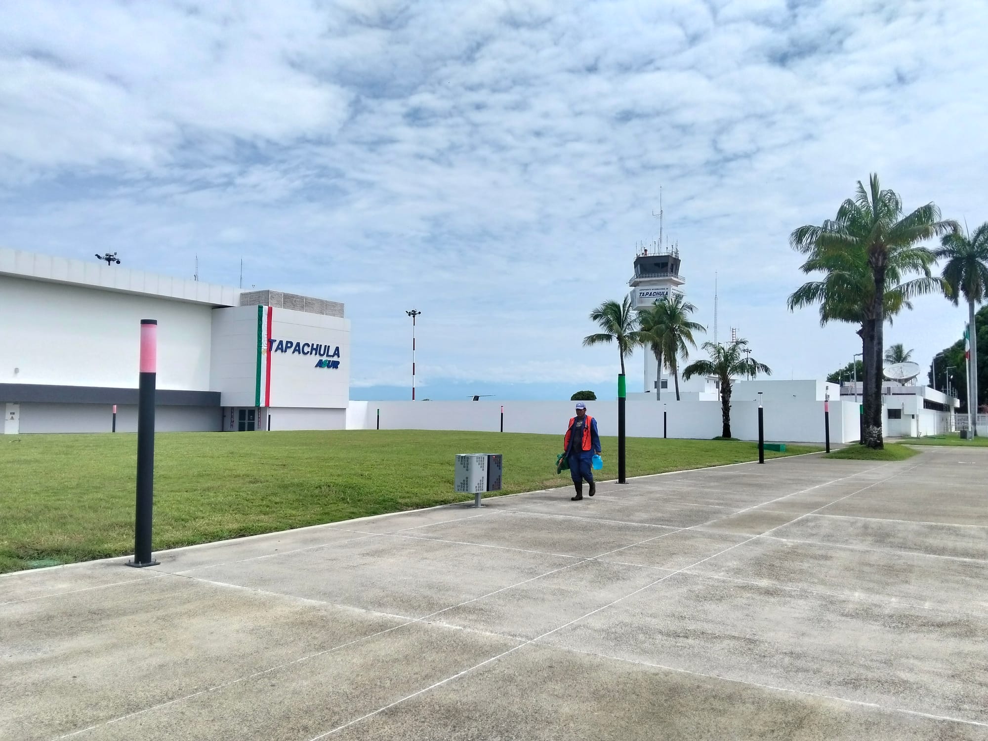 A man with an orange vest walks along the sidewalk in front of Tapachula airport with the control tower visible behind him