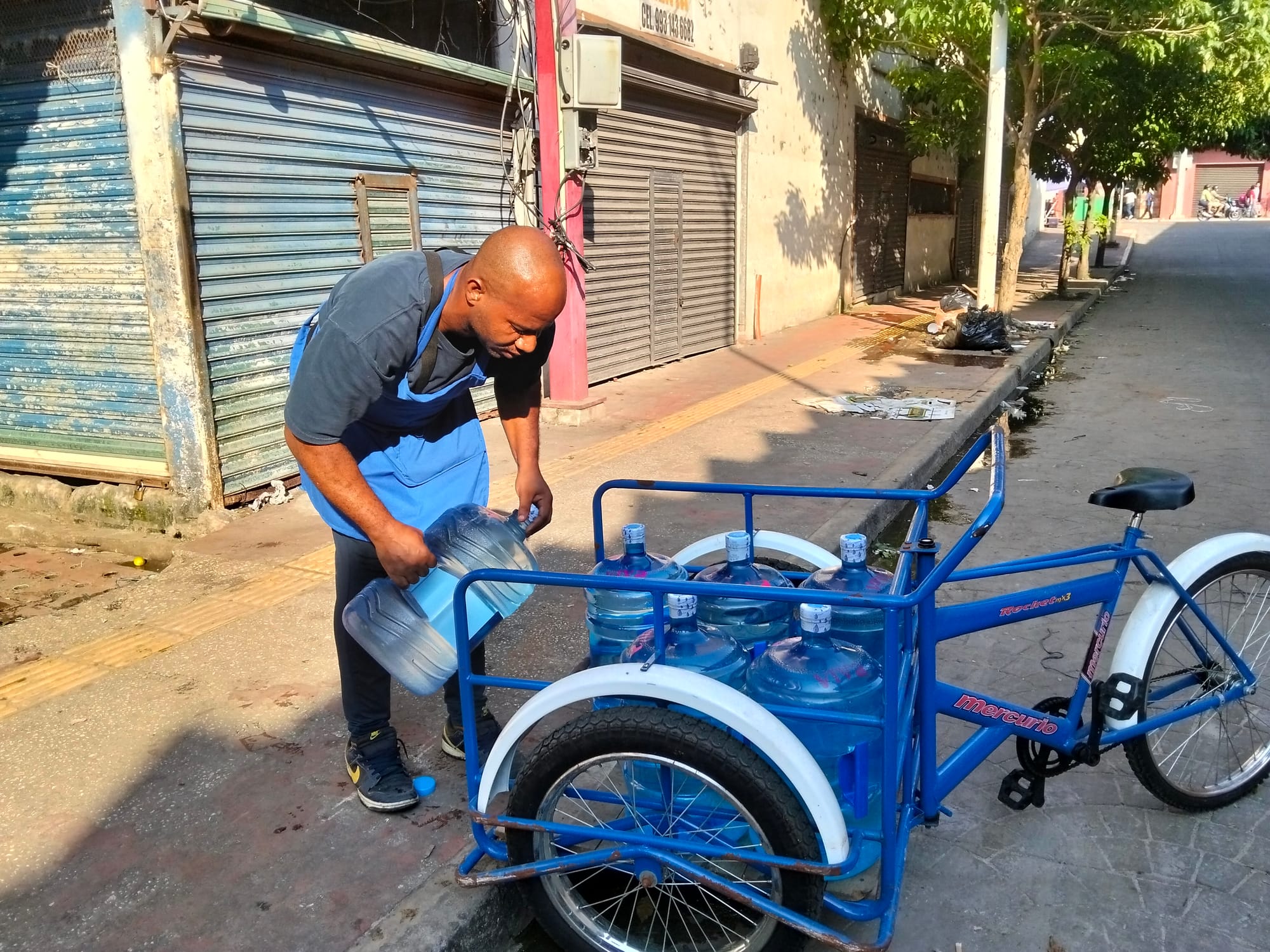 A man in a blue apron holds a jug of water next to a blue tricycle