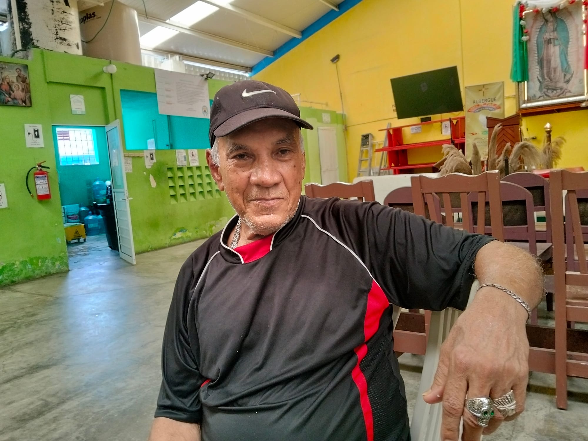 A man in a black and red shirt wearing rings sits in front of a green wall
