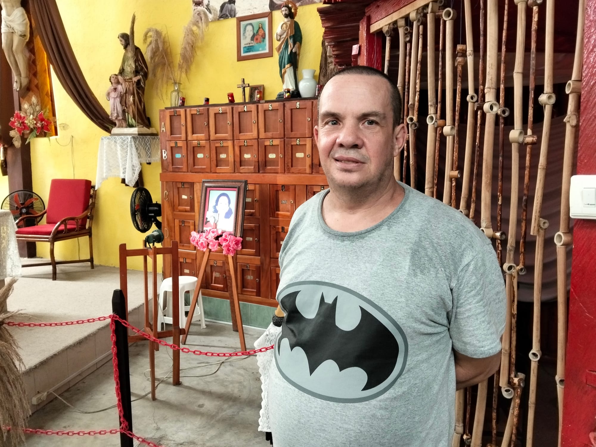 A man in a gray Batman shirt stands in front of a yellow wall