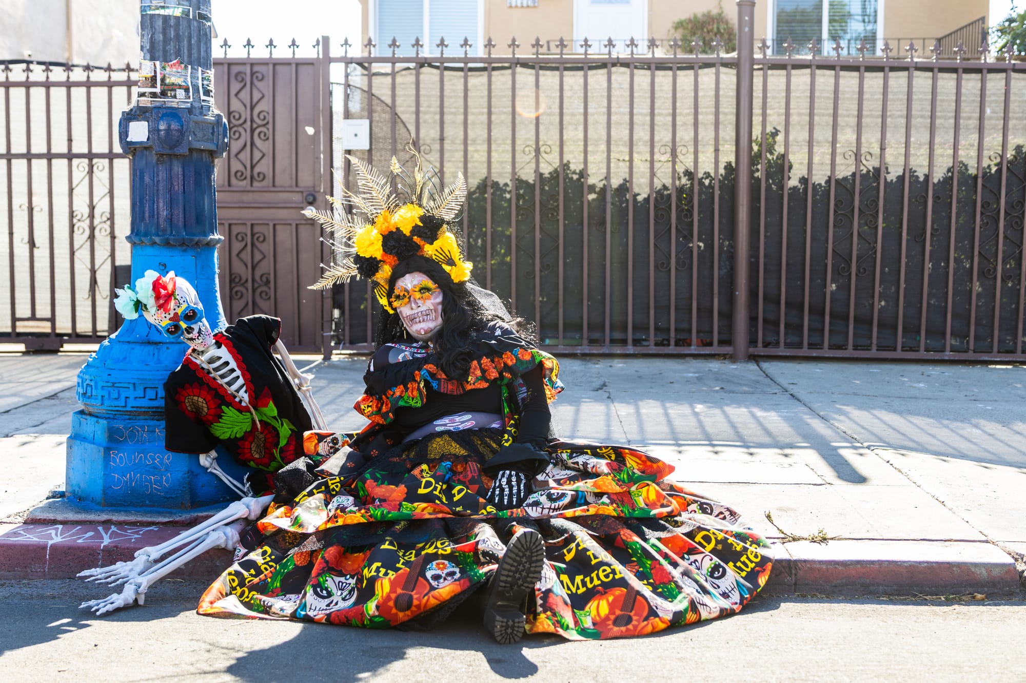A woman wearing a big dress sits next to a skeleton on the sidewalk.