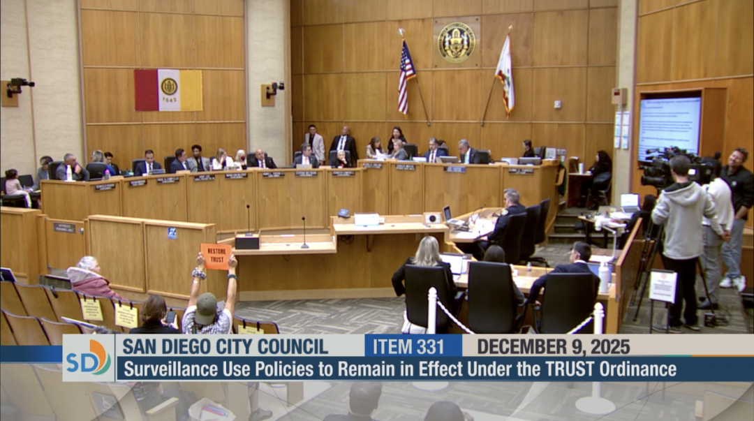 A screenshot of the livestream of the city council chambers with the members at their seats and one person in the audience holding up an anti-Flock sign