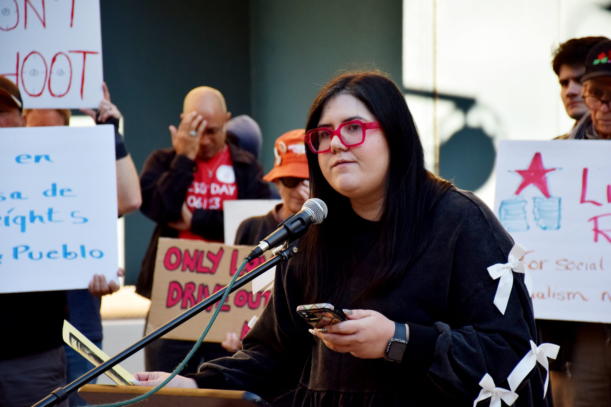 A person with pink framed glasses speaks outside at a podium in front of people holding signs.