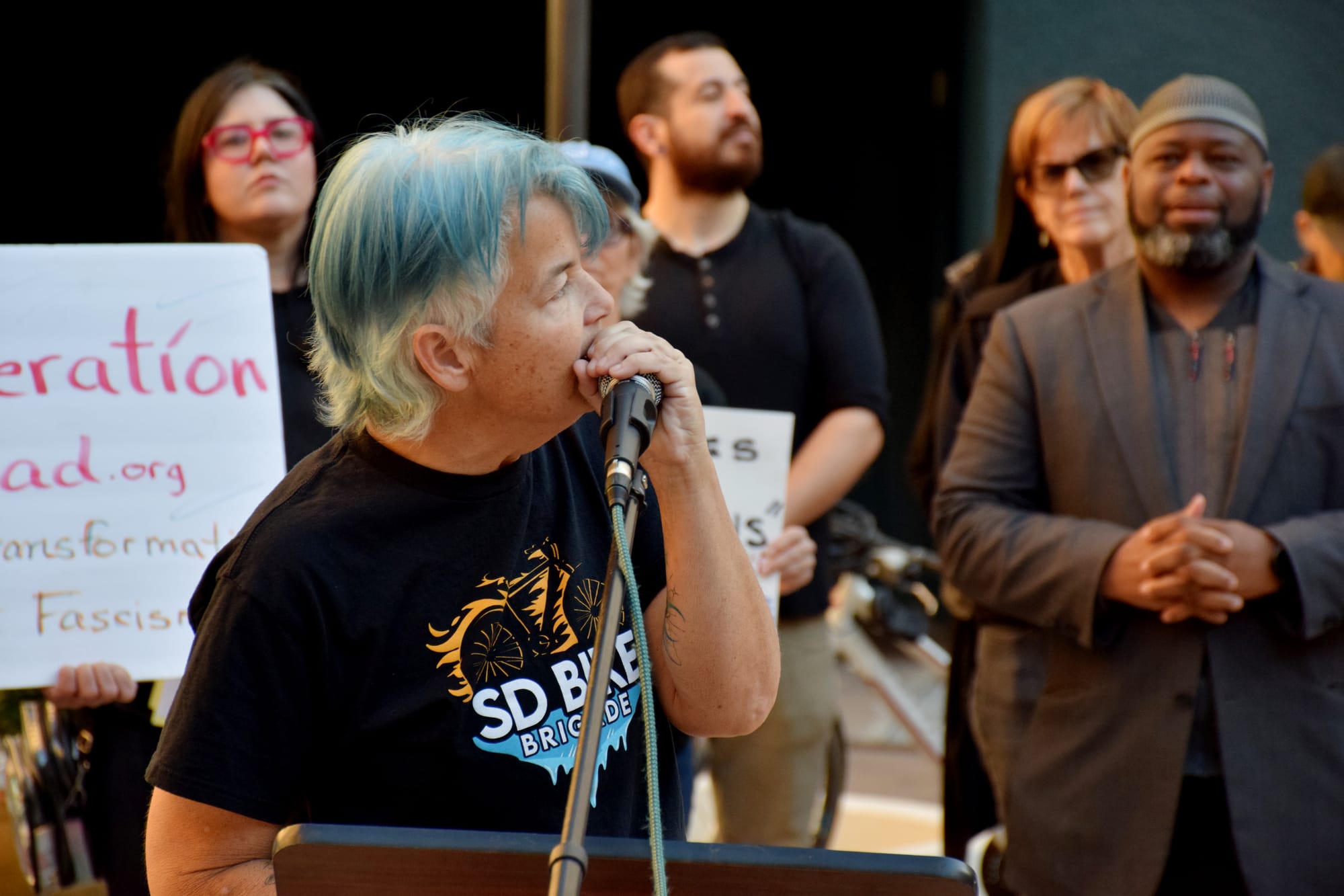 A person with blue hair speaks closely into a microphone with a crowd of people behind them.