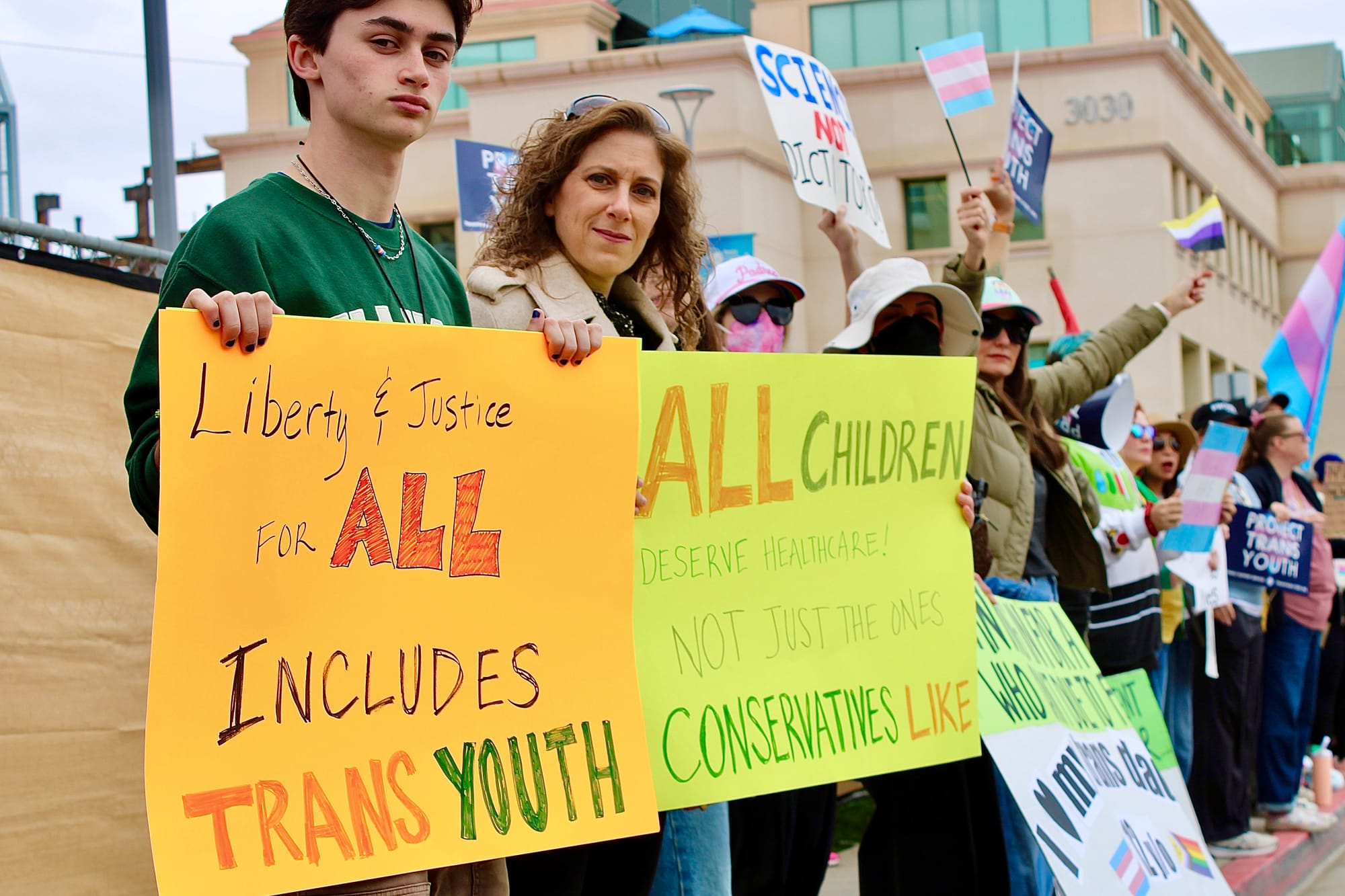 Two people hold signs in protest. They read, "Liberty and justice for all includes trans youth," and "All children deserve healthcare! Not just the ones conservatives like."