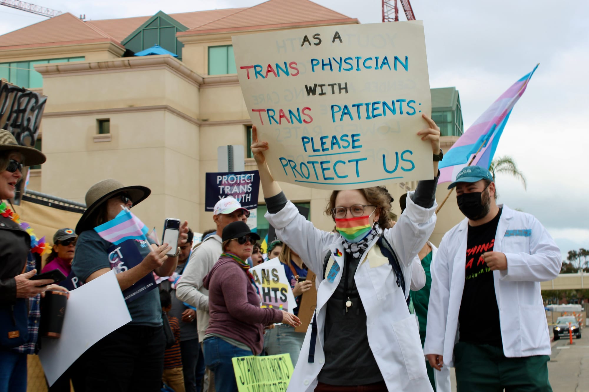 Health care workers protest with signs and transgender pride flags in white lab coats and march in a line adjacent to protestors.