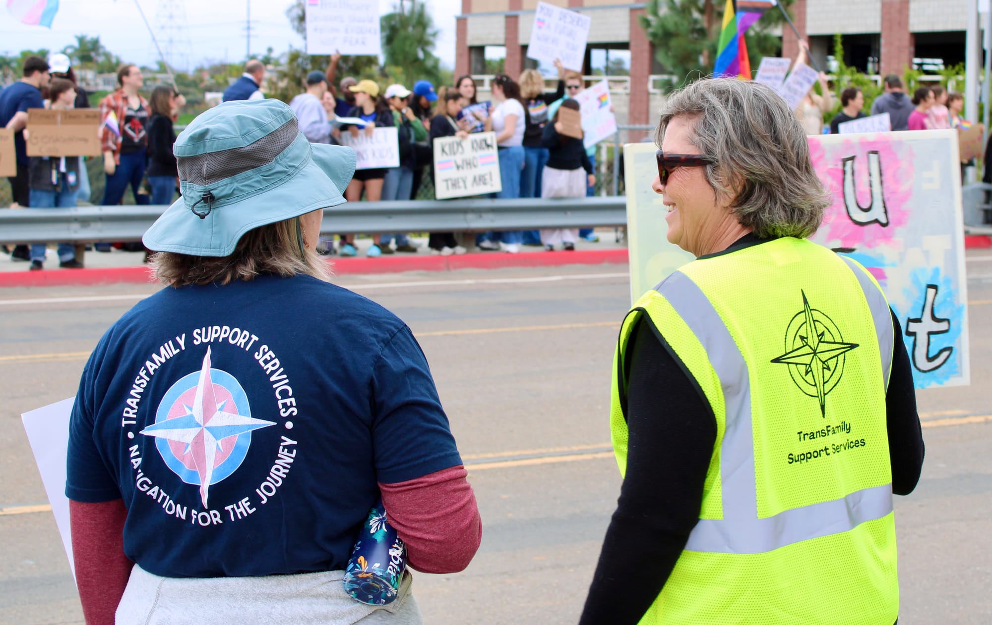 Two people in clothing that says "TransFamily Support Services" hold signs on the side of a road. The other side of the road is filled with protestors.