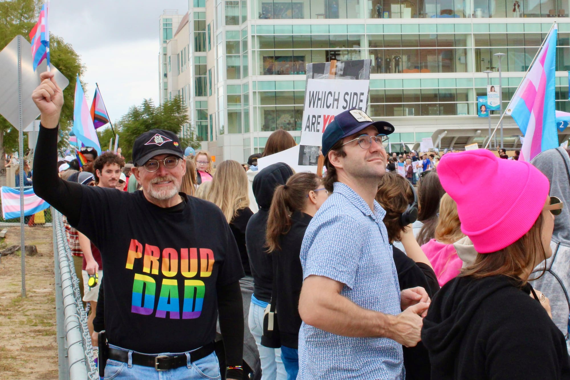 An older man wears a shirt that reads "Proud Dad," as he waves a transgender pride flag in a crowd of protestors.
