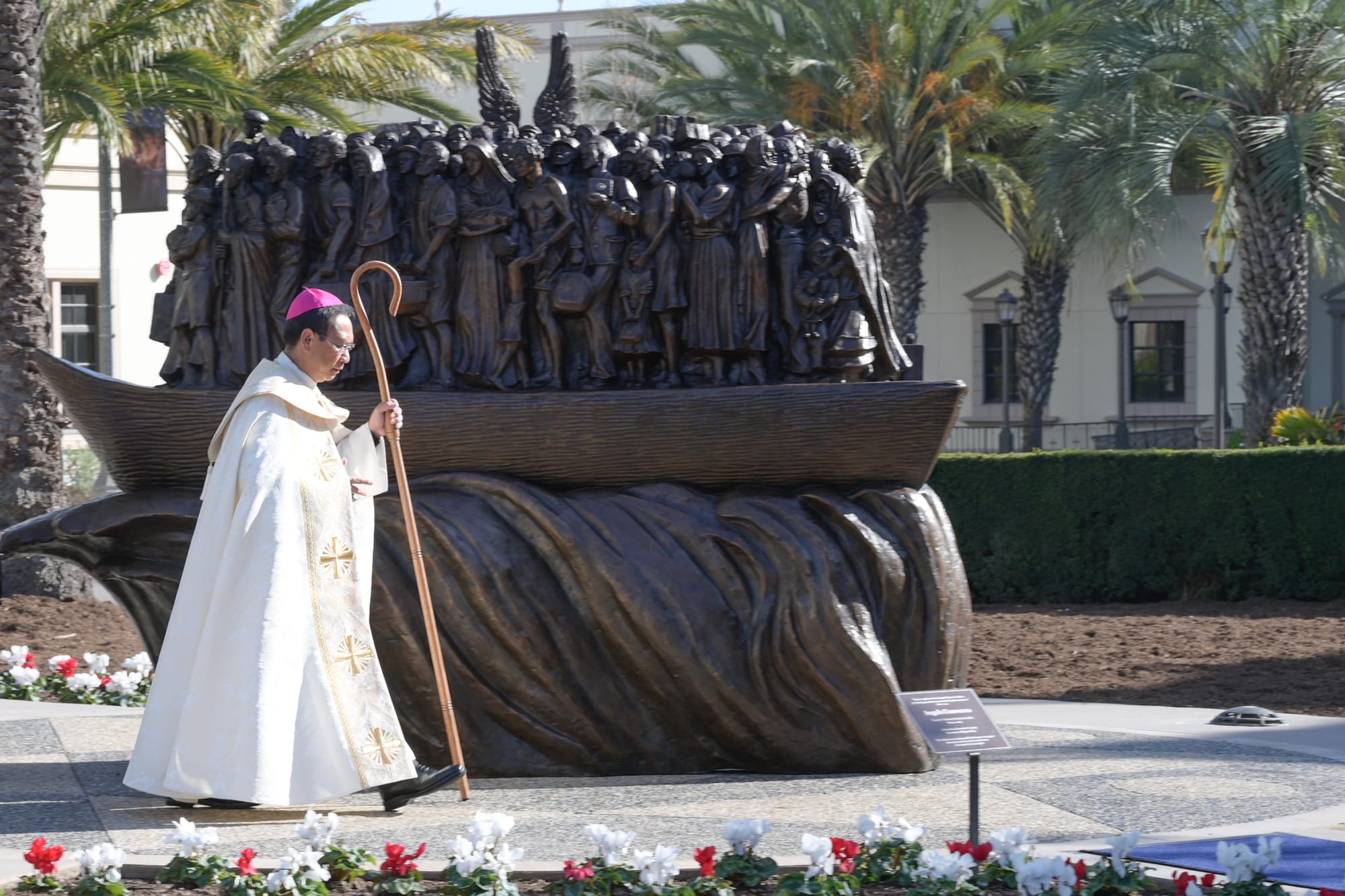 A man in bishop's robes walks next to a bronze statue of migrants on a boat