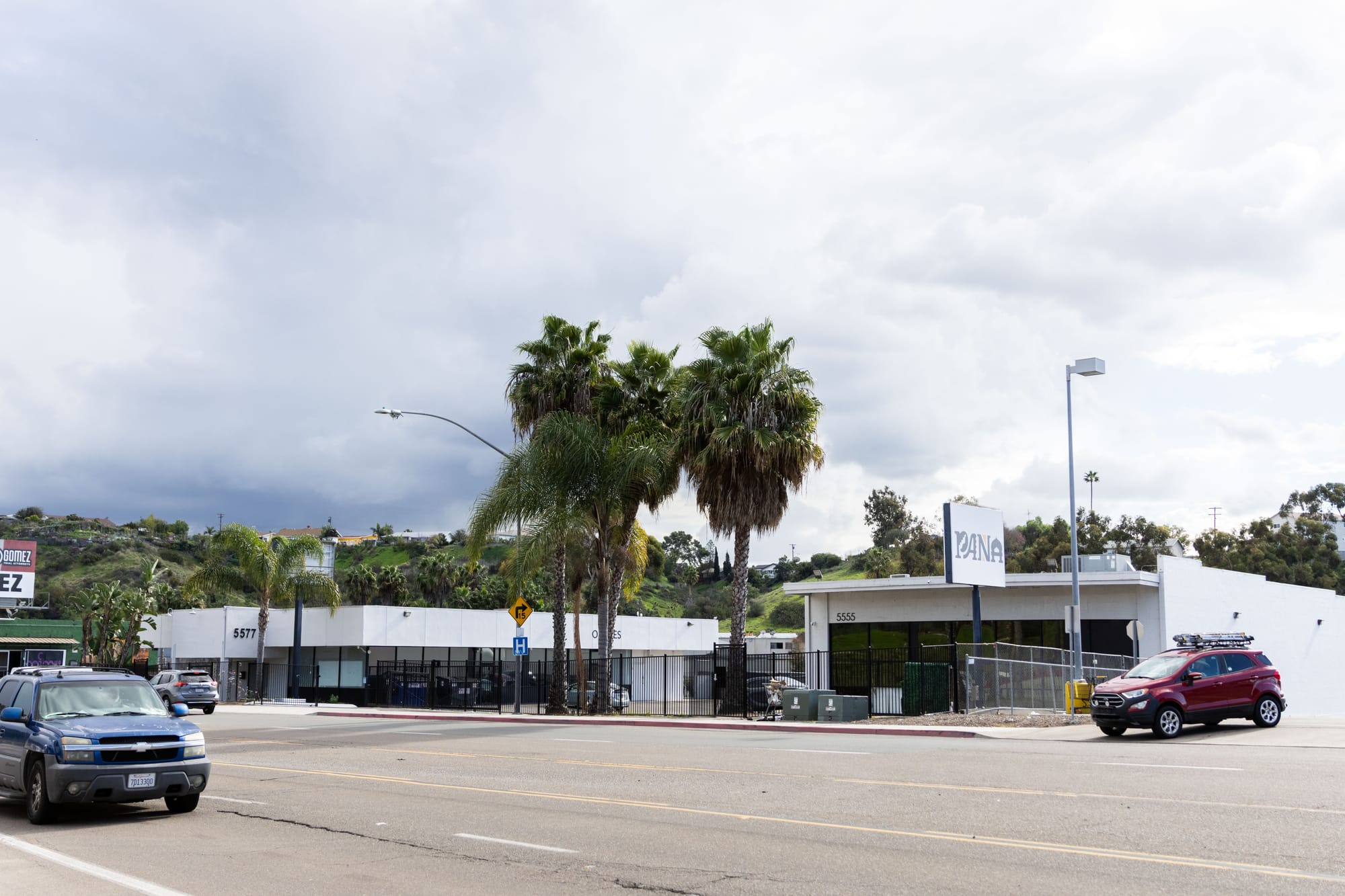 A building across the street with several trees, street lights, cars and a sign in front of it.