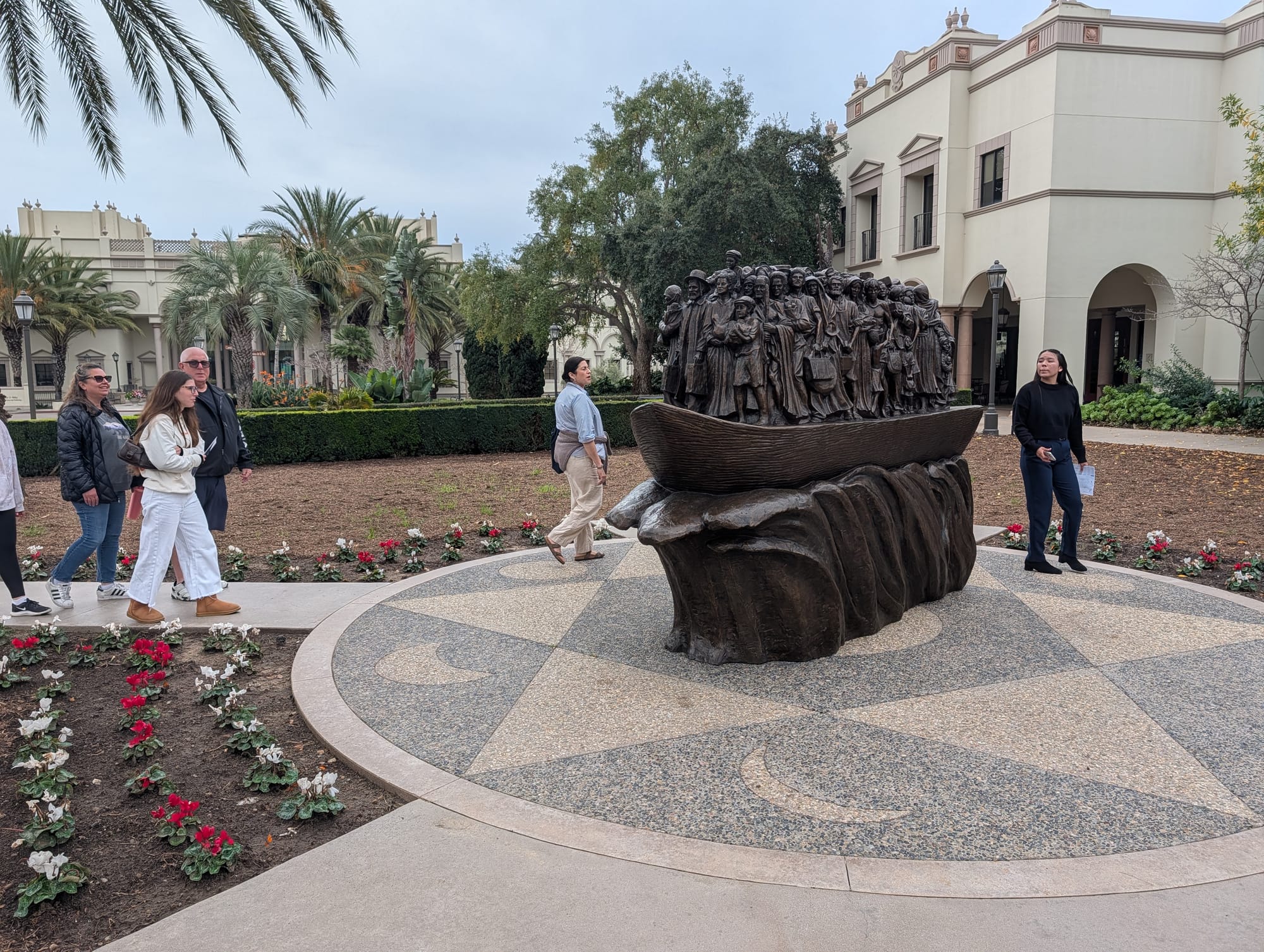 A woman in black clothing and a woman in a blue shirt and khaki pants stand near a bronze statue of migrants on a boat with university buildings in the background