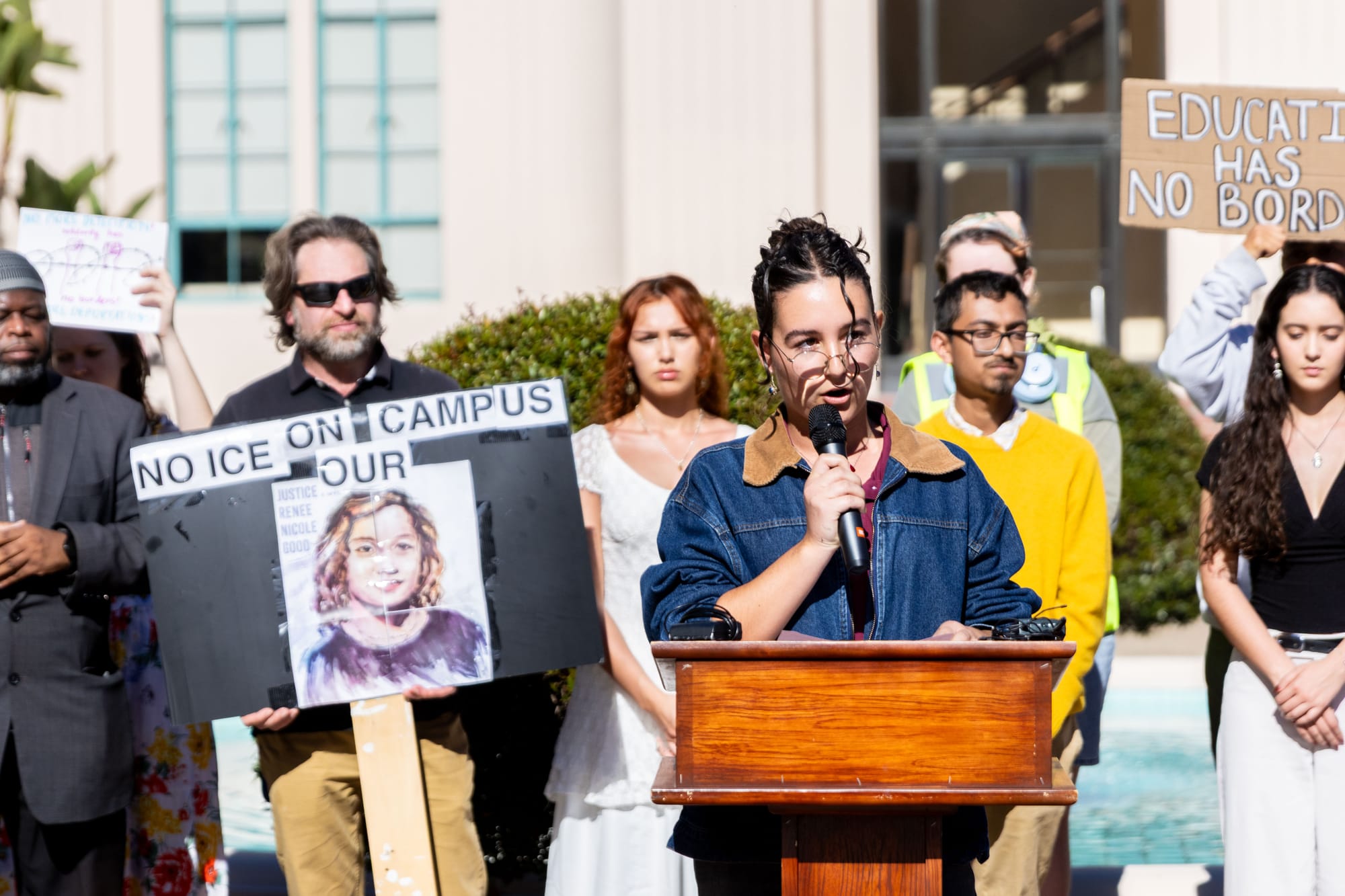A woman holding a microphone stands at a podium with people behind her holding up signs.