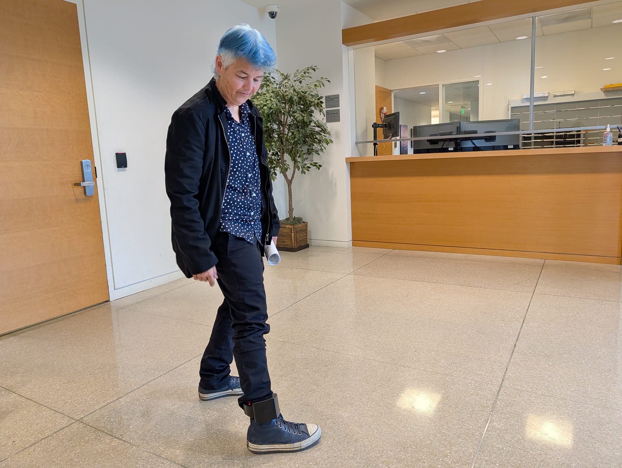 A woman with blue hair looks down at an ankle monitor on her leg in the lobby of an office