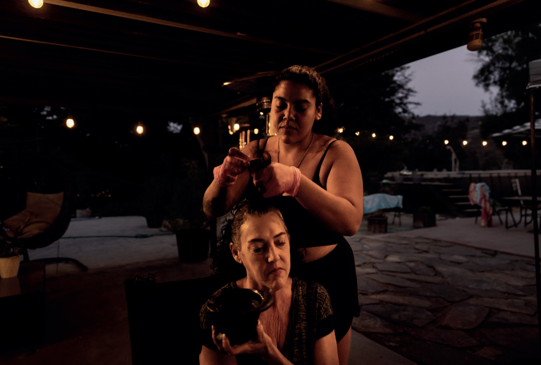 Picture of two women outside, one sitting have her hair dyed by the other.