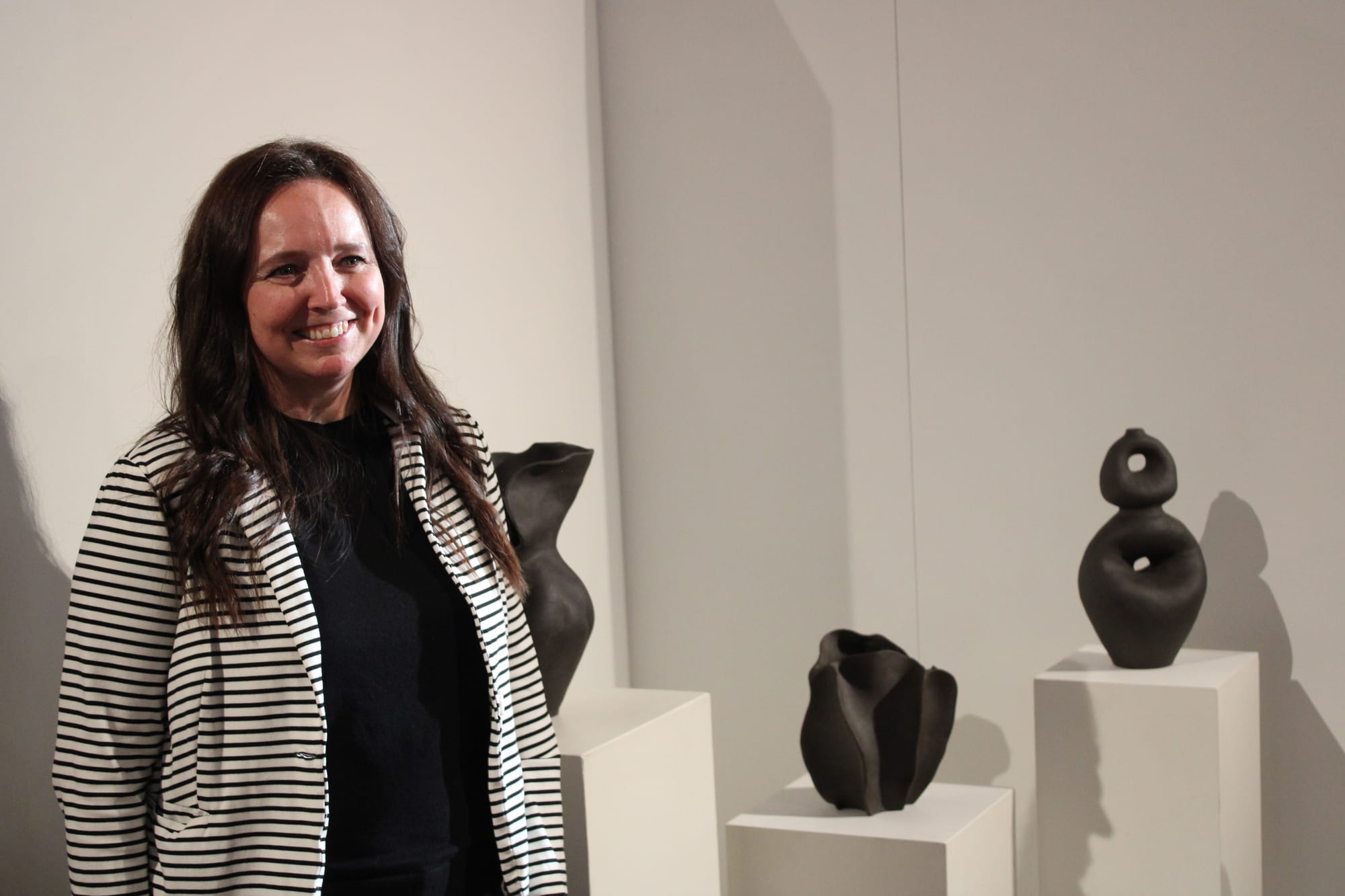 A woman in a black and white striped jacket smiles and stands beside dark gray pottery on stands.