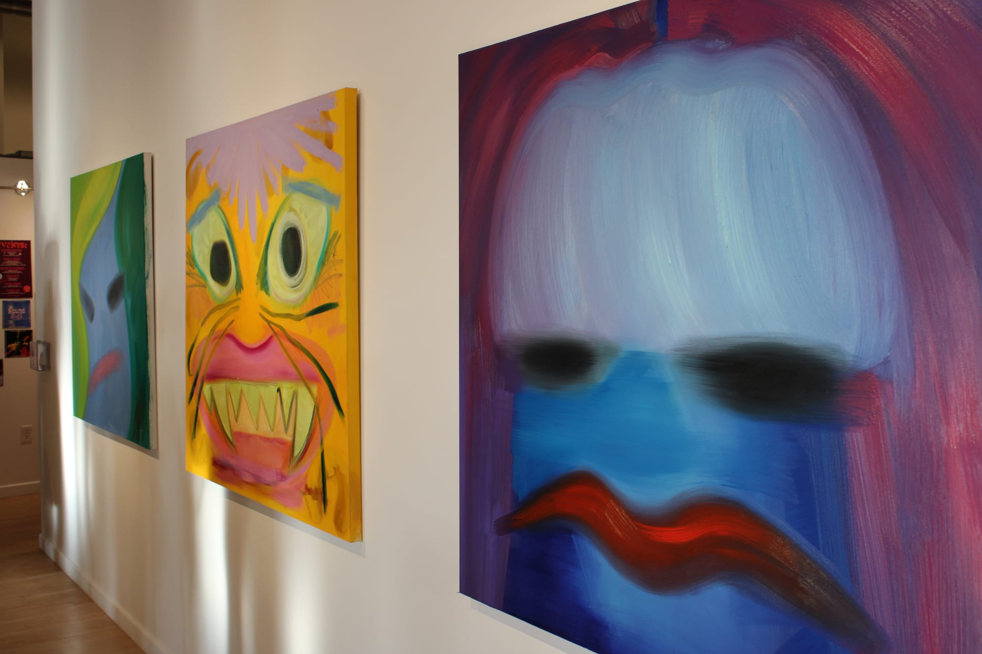 Three colorful oil paintings that look like abstract faces or masks are hung in a line on a white wall.