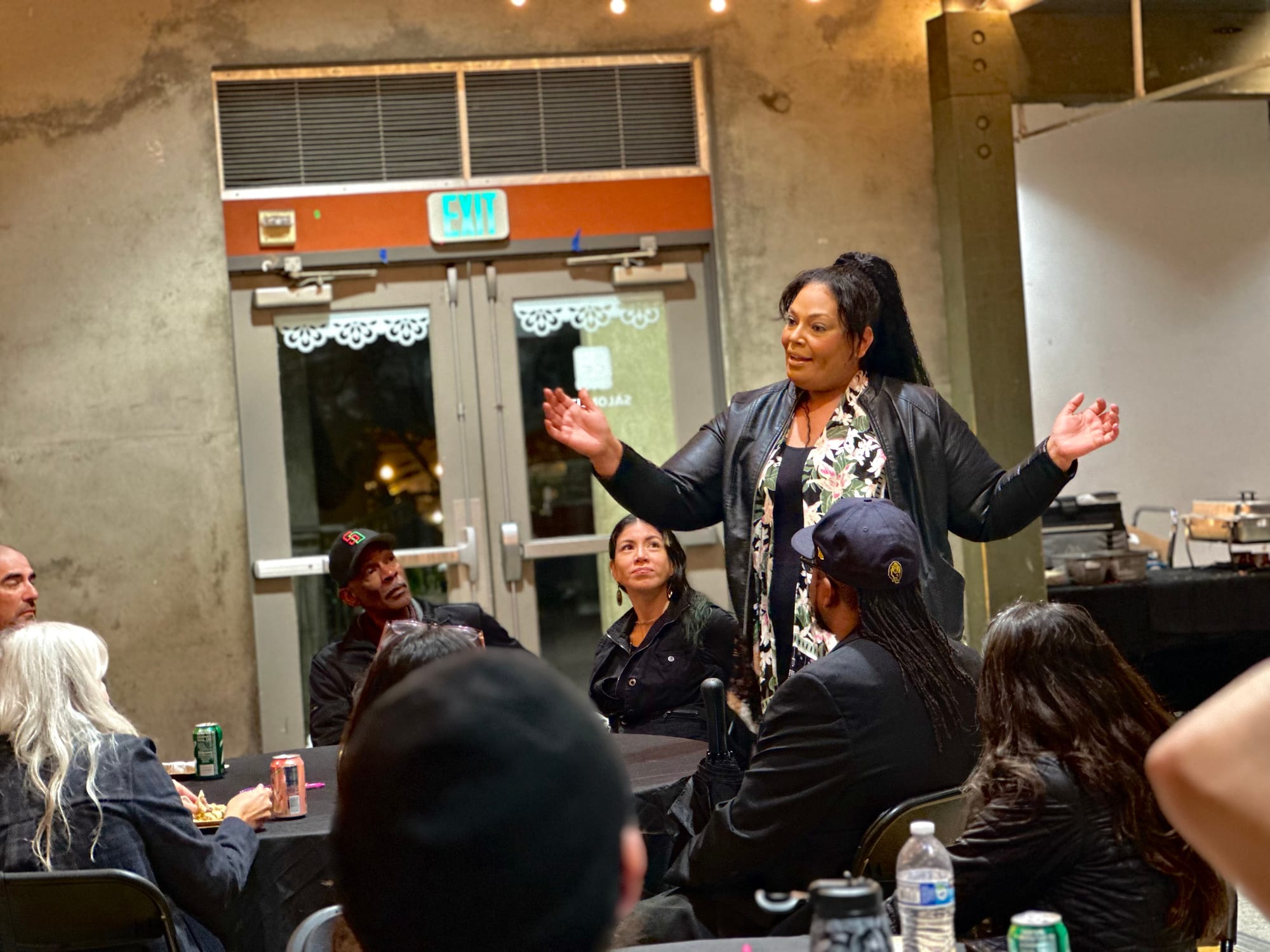 A woman in a black leather jacket stands up with her arms out and speaks to a crowd of people who are sitting down.