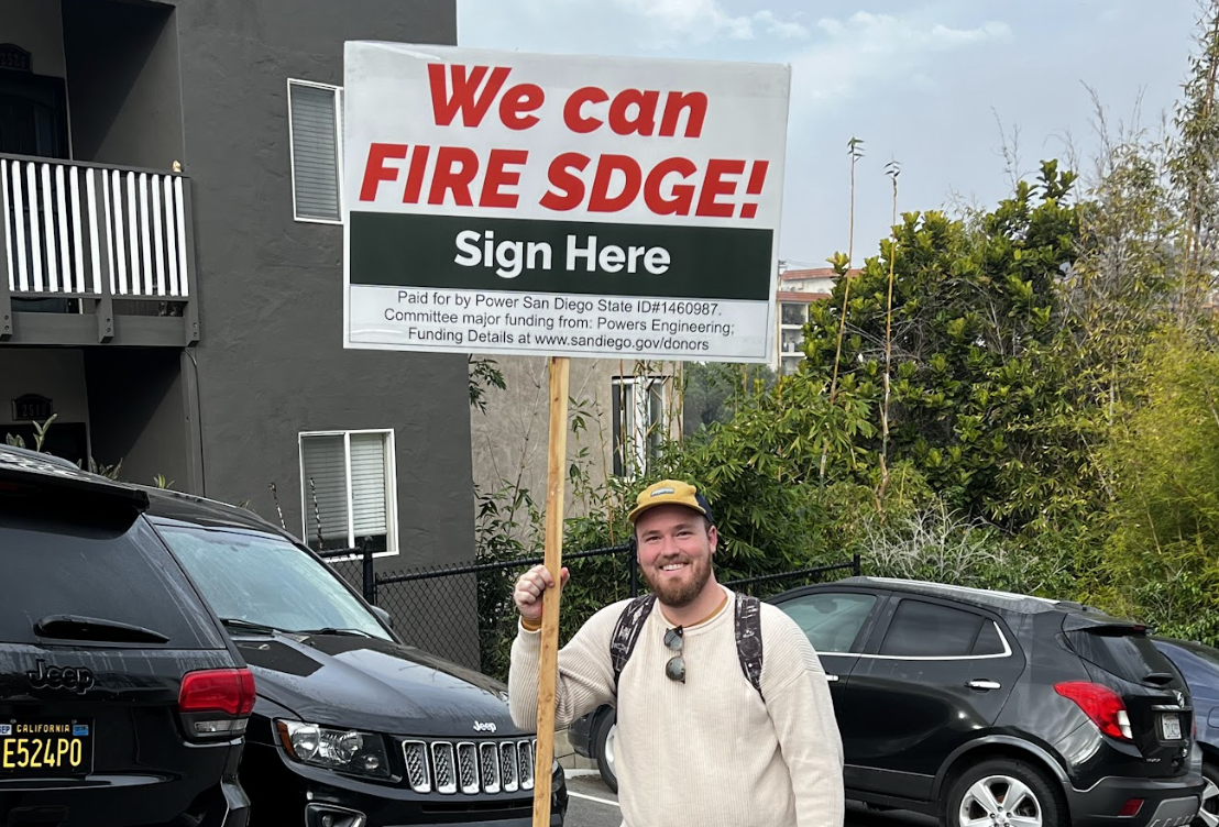 A man in a hat holds a large, tall sign that reads, "We can fire SDGE."