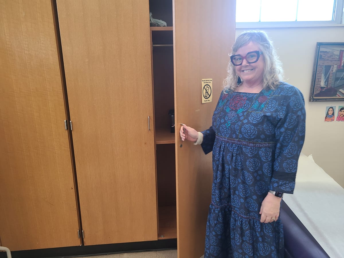 A woman in a blue floral dress and glasses opens a classroom storage closet door.