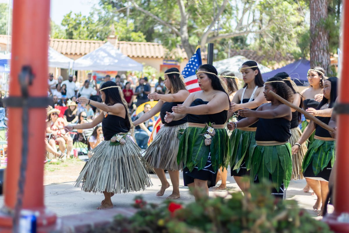 Dancers in grass and leaf skirts perform on a stage during an outdoor event with people in the background