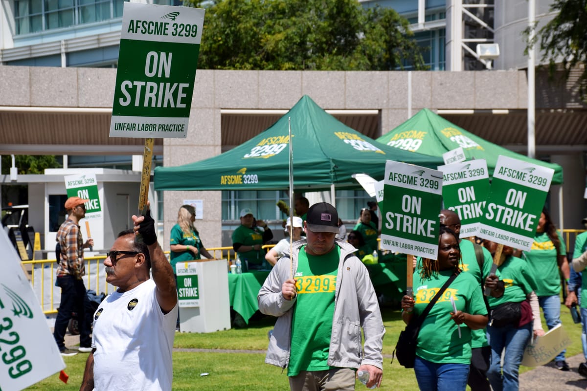 People outside walk holding green and white picket signs that say "AFSCME 3299 On Strike Unfair Labor Practices" on the grass outside of a hospital