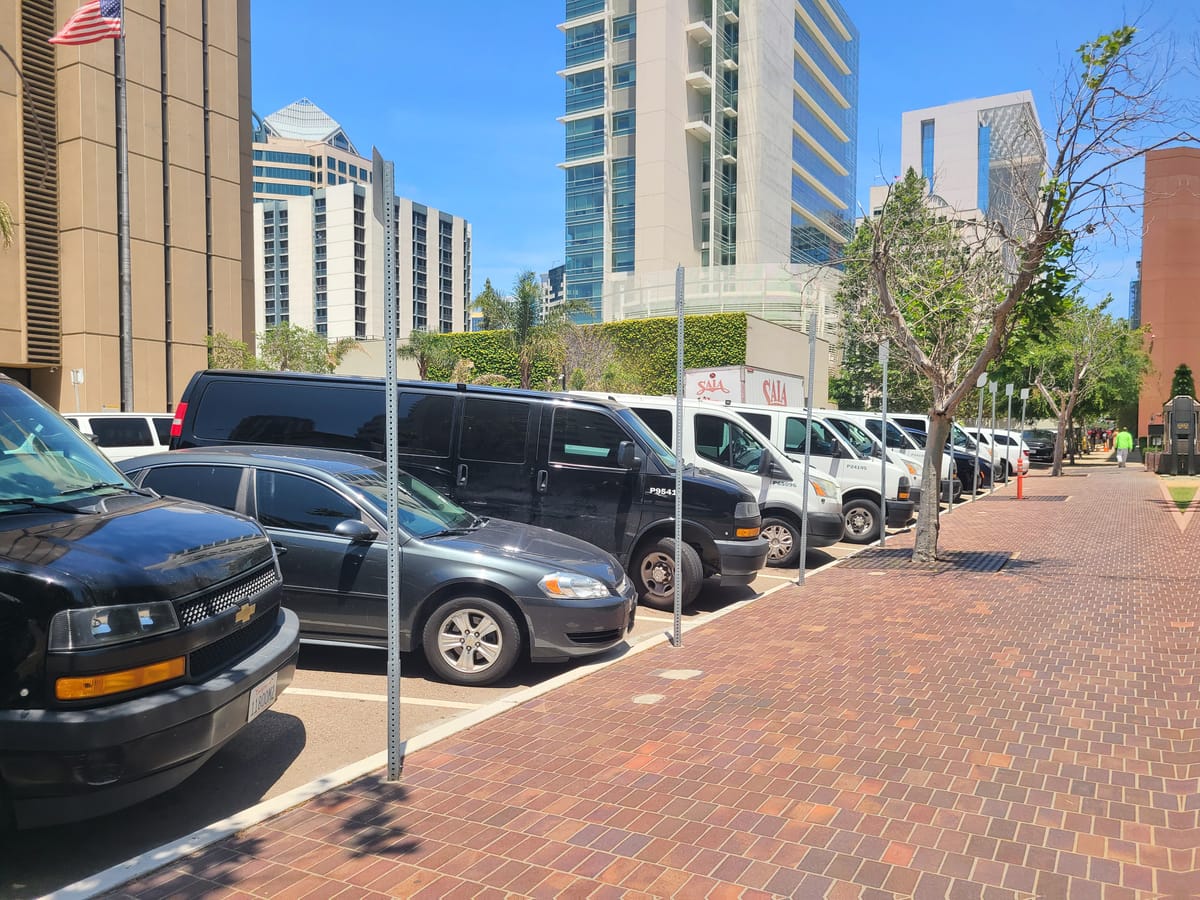 A black sedan, a black van and several white sedans are parked in a row in front of a brick sidewalk.