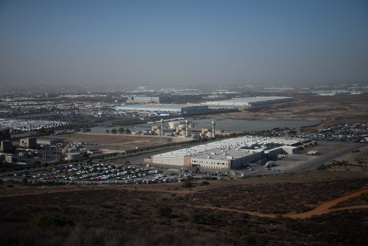 A prison-like building sits at the bottom of a hill with an Amazon warehouse in the background.