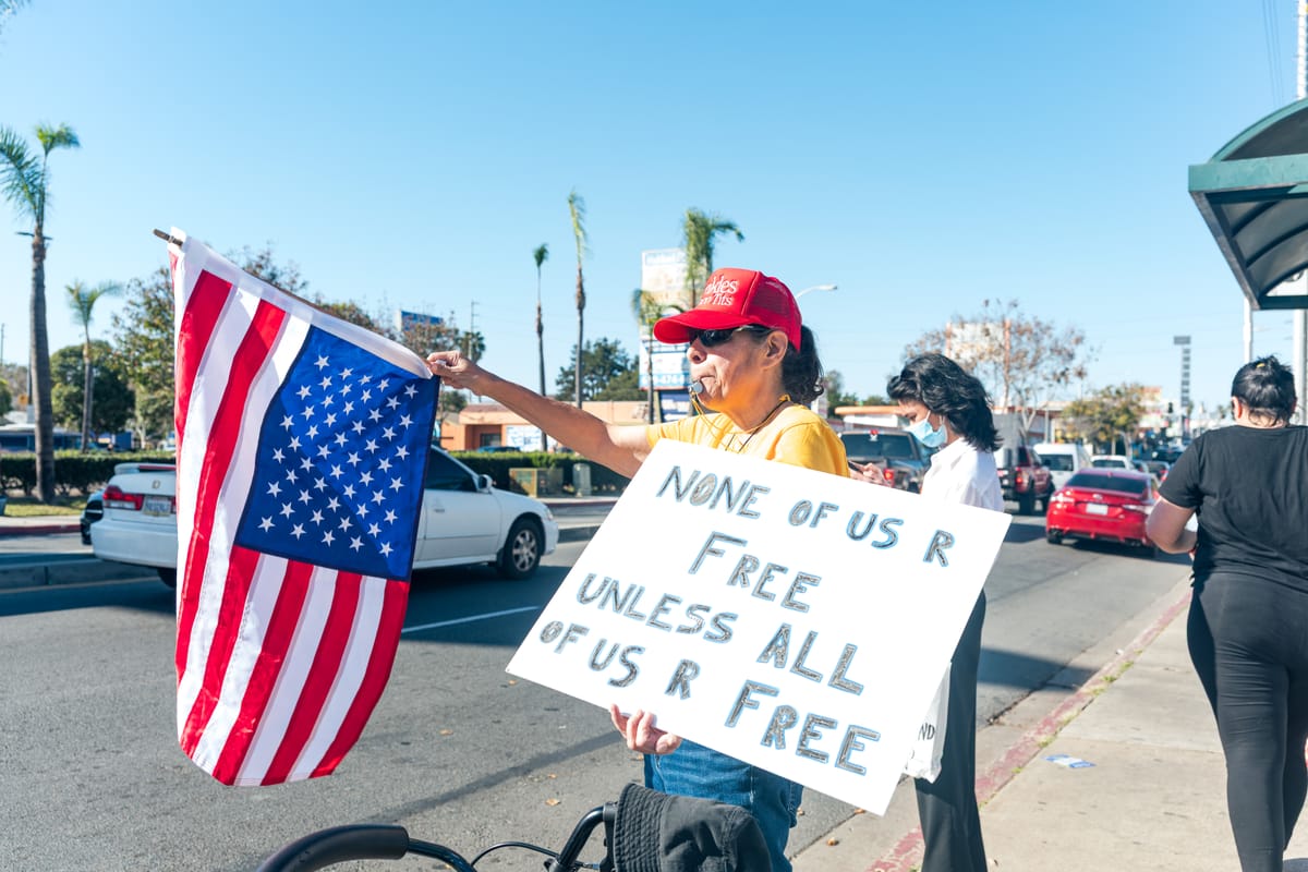 A person in a red had holding an American flag and holding a sign that says "None of us R Free Unless All of Us R Free"